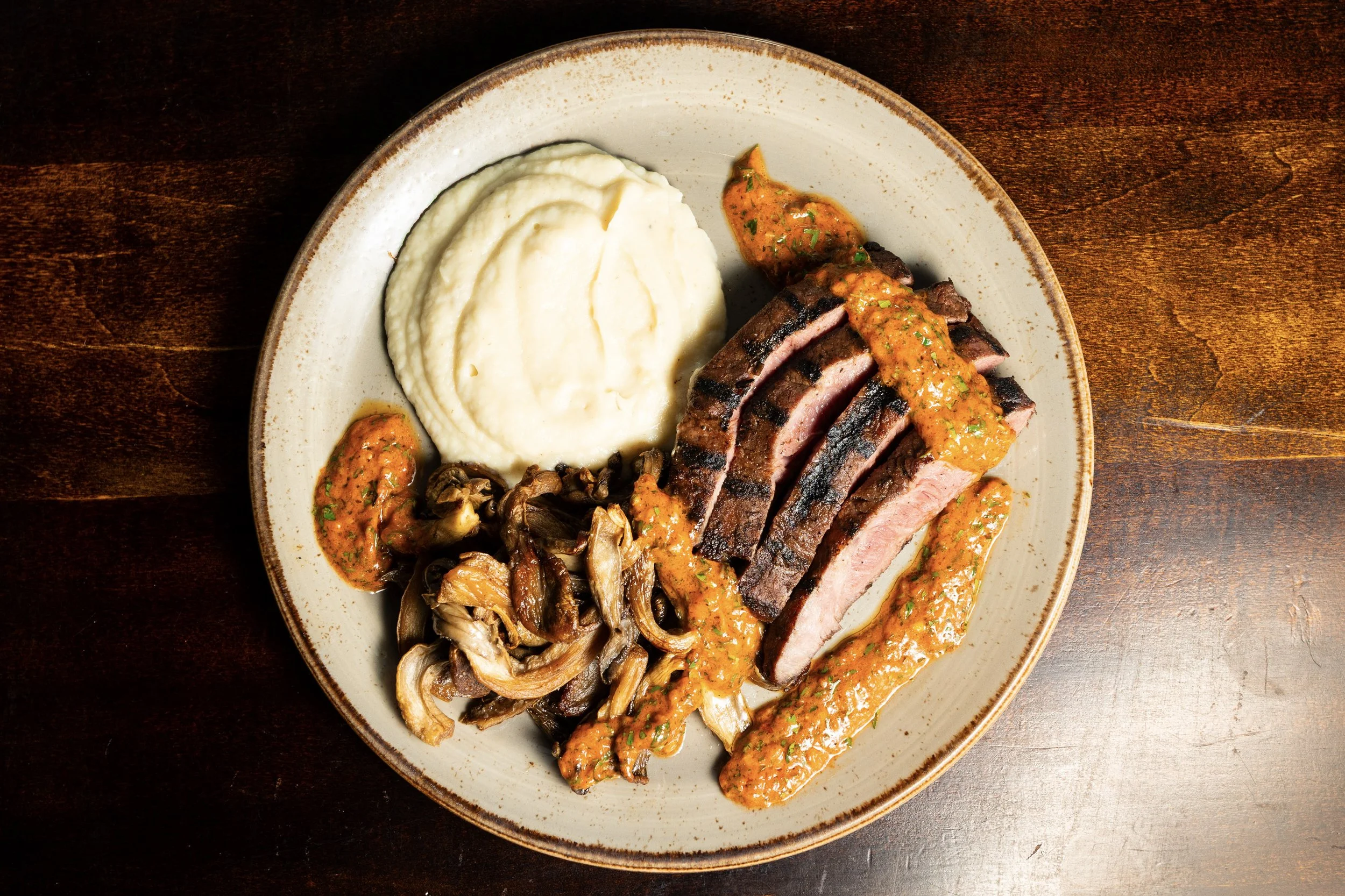 Plate of sliced grilled steak with sauce, mashed potatoes, and sautéed mushrooms on a wooden table.