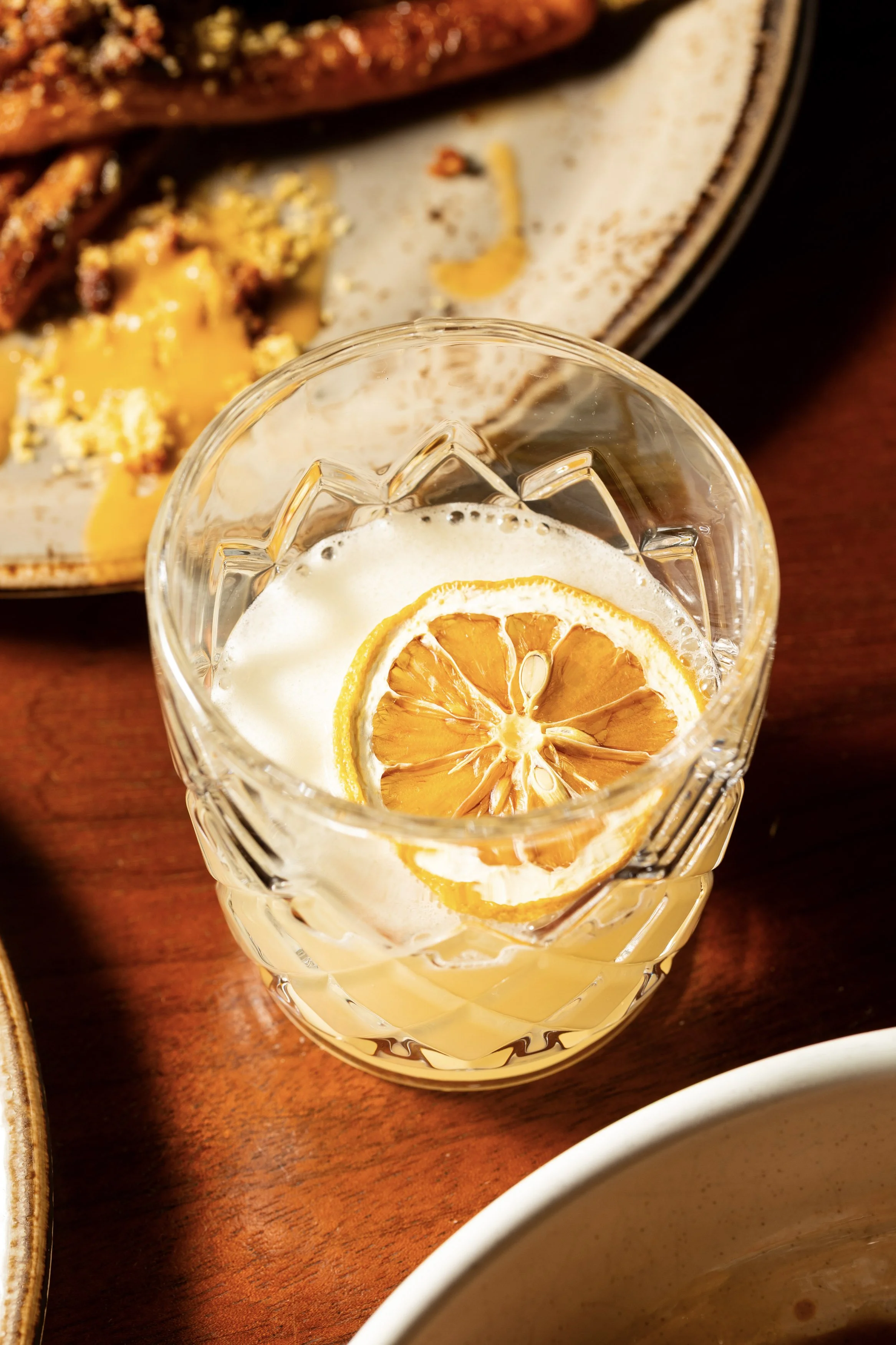 A glass of lemonade with lemon slices garnishing the drink on a wooden table. In the background, part of a plate with breakfast food such as eggs and bacon is visible.