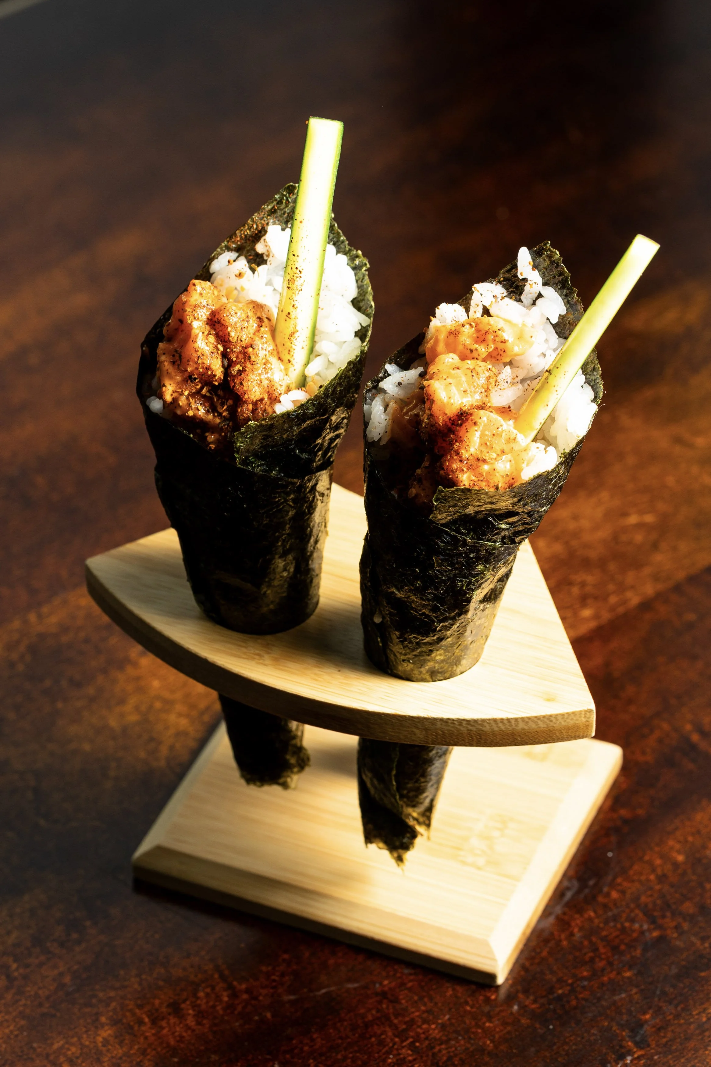Two sushi burritos in seaweed wraps filled with rice, spicy seafood, and cucumber slices, placed on a wooden serving stand.