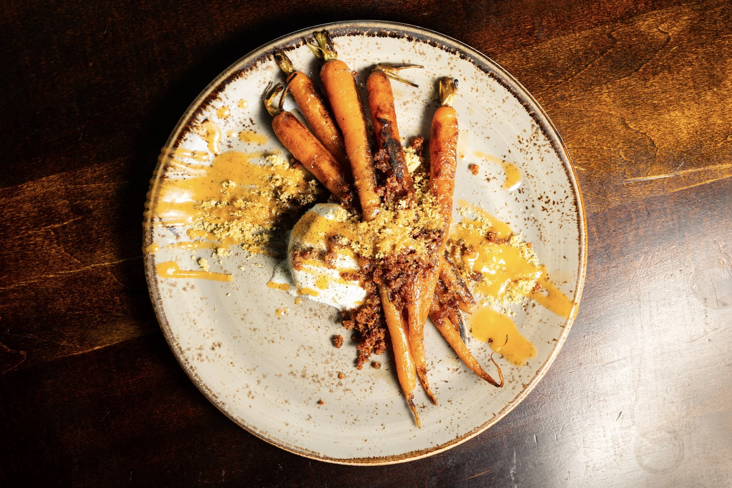 Roasted carrots on a plate garnished with crumbled cheese, drizzled with sauce, and served with a dollop of creamy white dip, on a dark wooden surface.