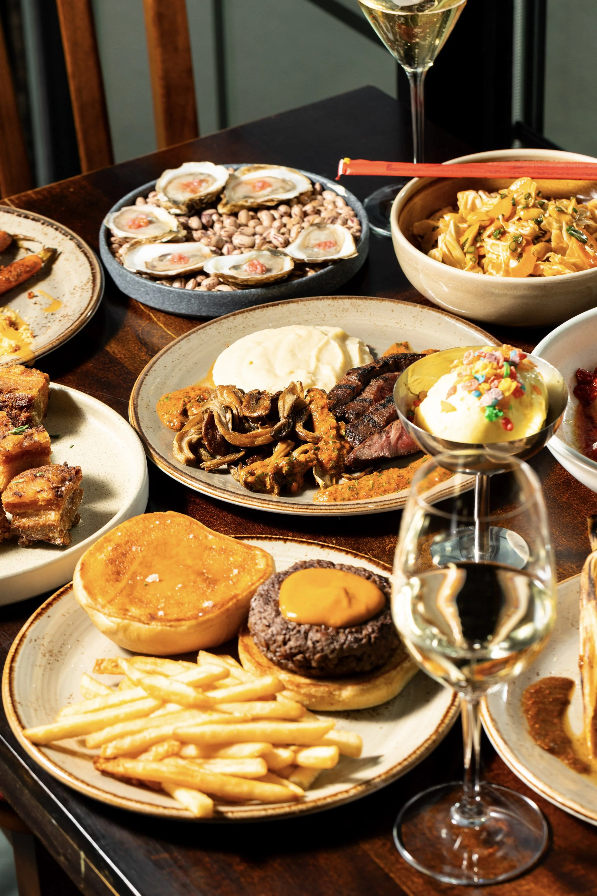 A table full of assorted dishes including oysters, pasta, stew, and burger sliders, with wine and dessert such as ice cream and cake.