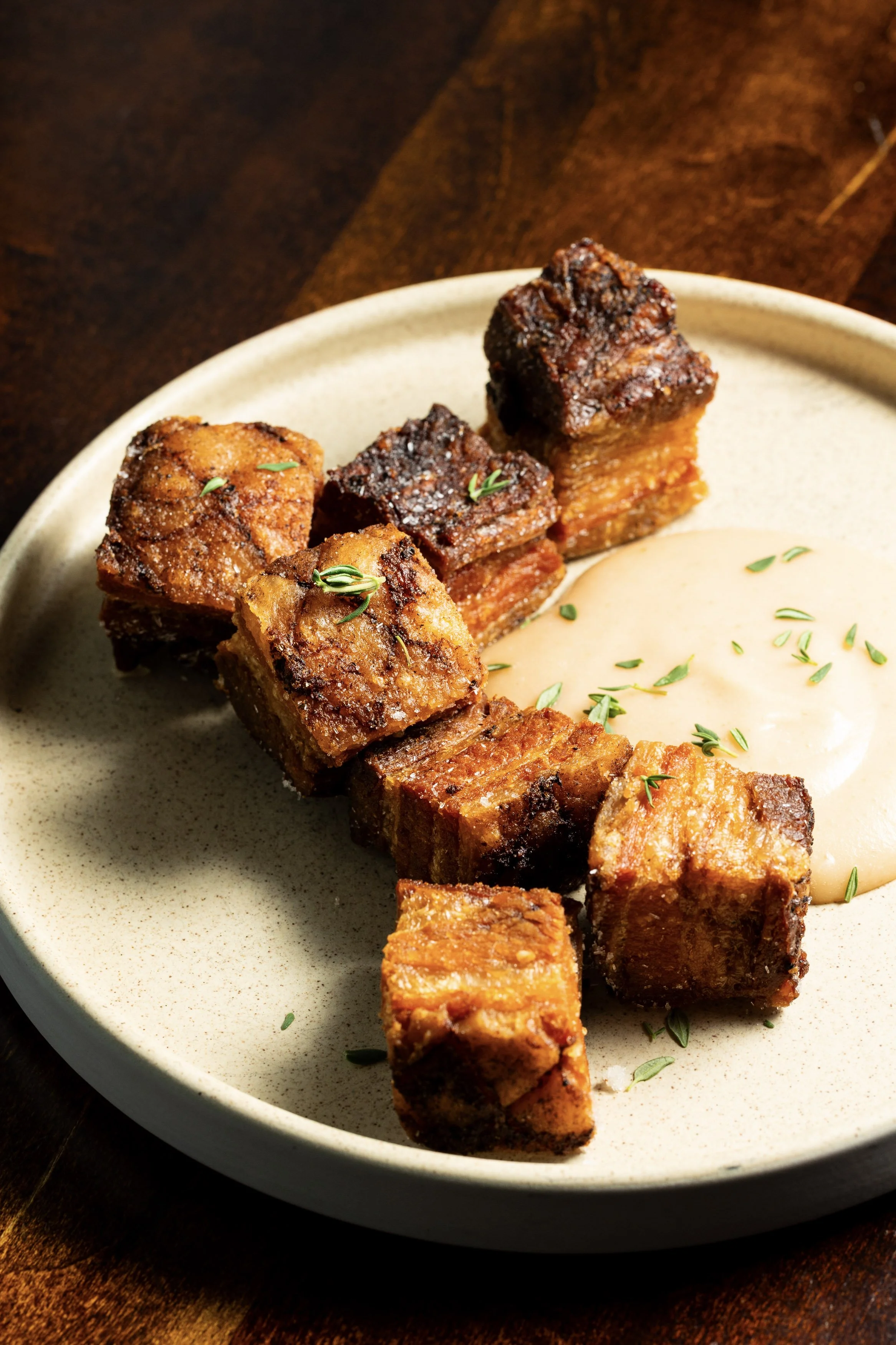 Fried pork belly cubes with a side of creamy sauce garnished with herbs on a beige plate