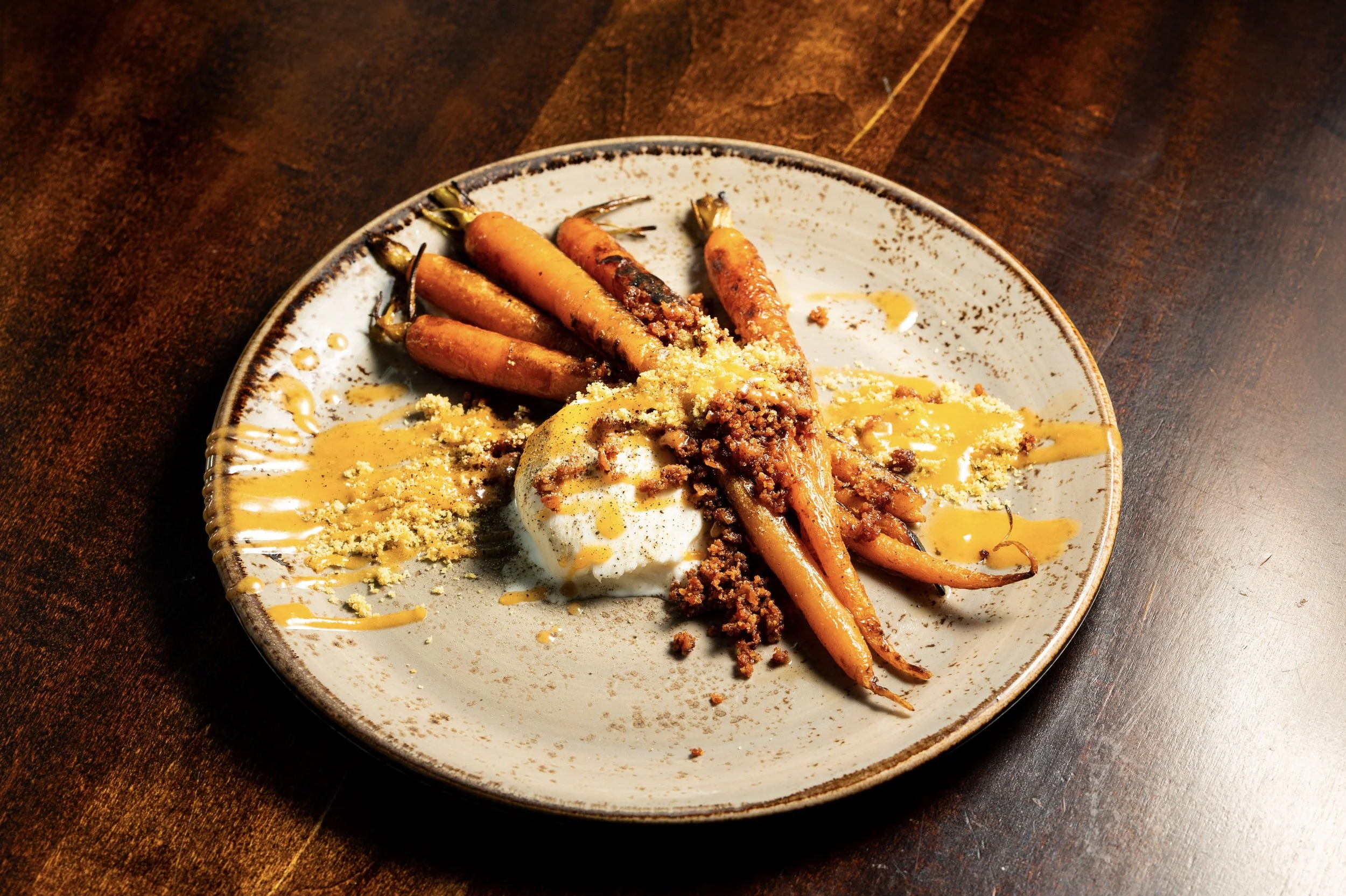 A plate of cooked carrots topped with crumbled crumbs and sauce, served with a dollop of sour cream and drizzled with a yellow sauce, on a dark wooden table.