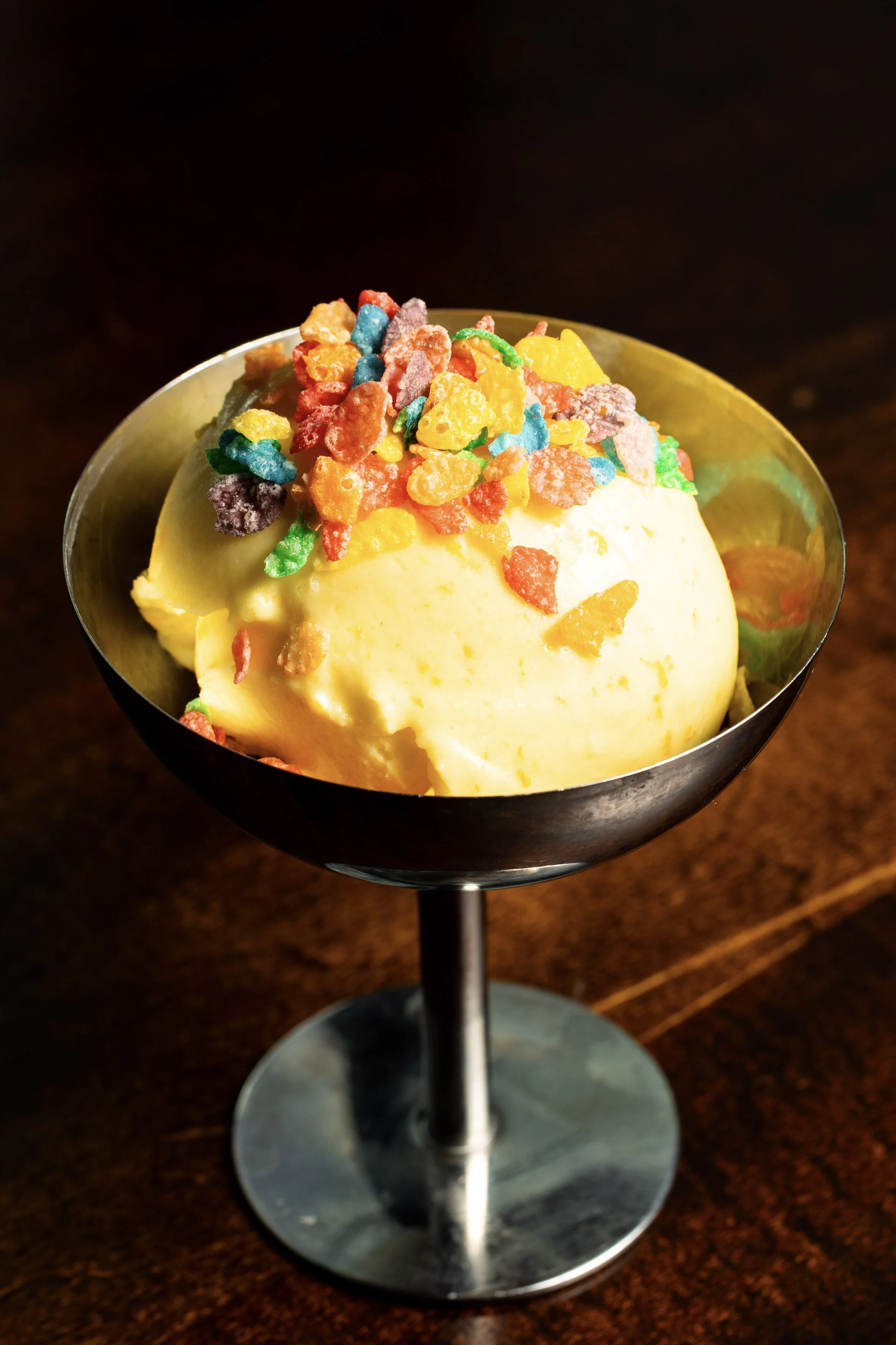 Scoops of yellow ice cream topped with colorful cereal pieces in a metal dessert dish on a wooden surface.