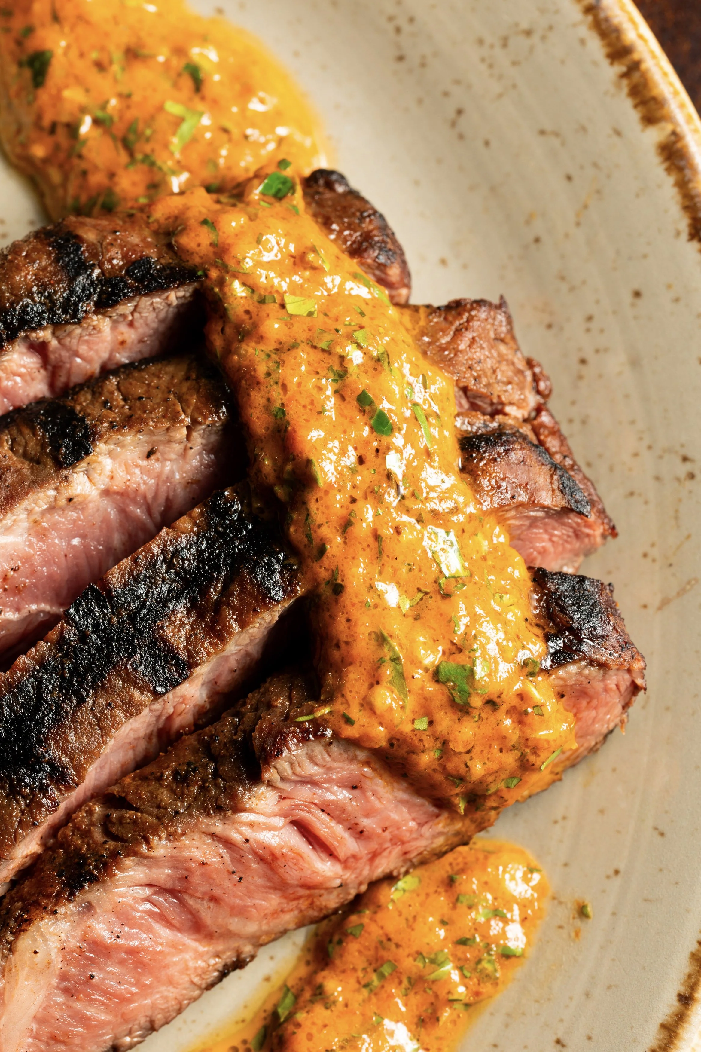 A close-up of sliced grilled steak topped with orange sauce and garnished with chopped herbs on a ceramic plate.