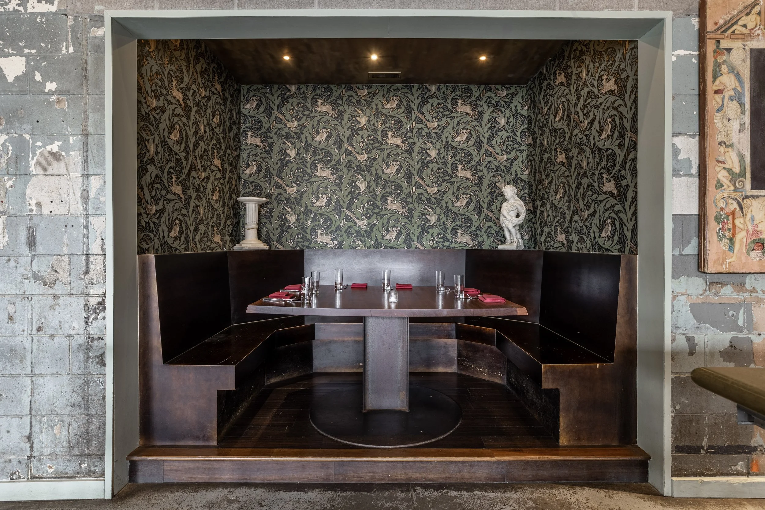 A cozy restaurant booth with dark wooden seating around a rectangular table, set with glassware and red napkins. The booth's back wall features green and beige patterned wallpaper, and two decorative statues are placed on small pedestals on either side of the corner.