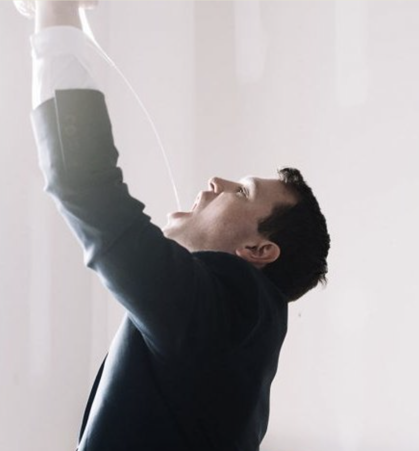 A man wearing a dark blazer and white shirt holds a water bottle above his head, pouring water into his mouth.