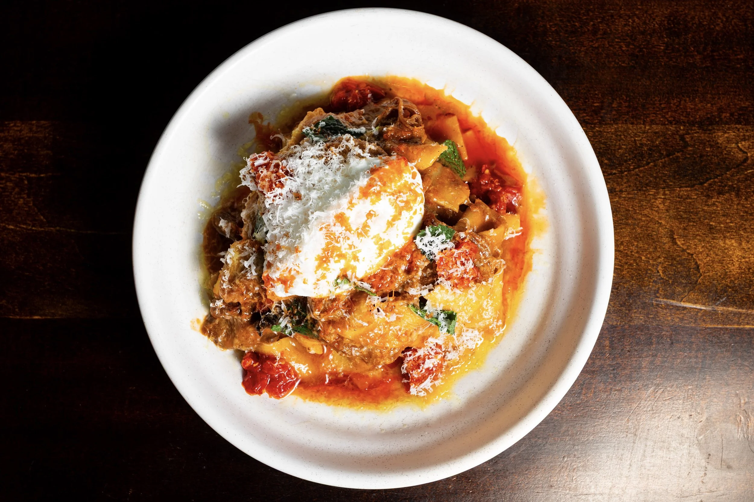 A bowl of beef stew topped with shredded cheese and a poached egg, served on a wooden table.