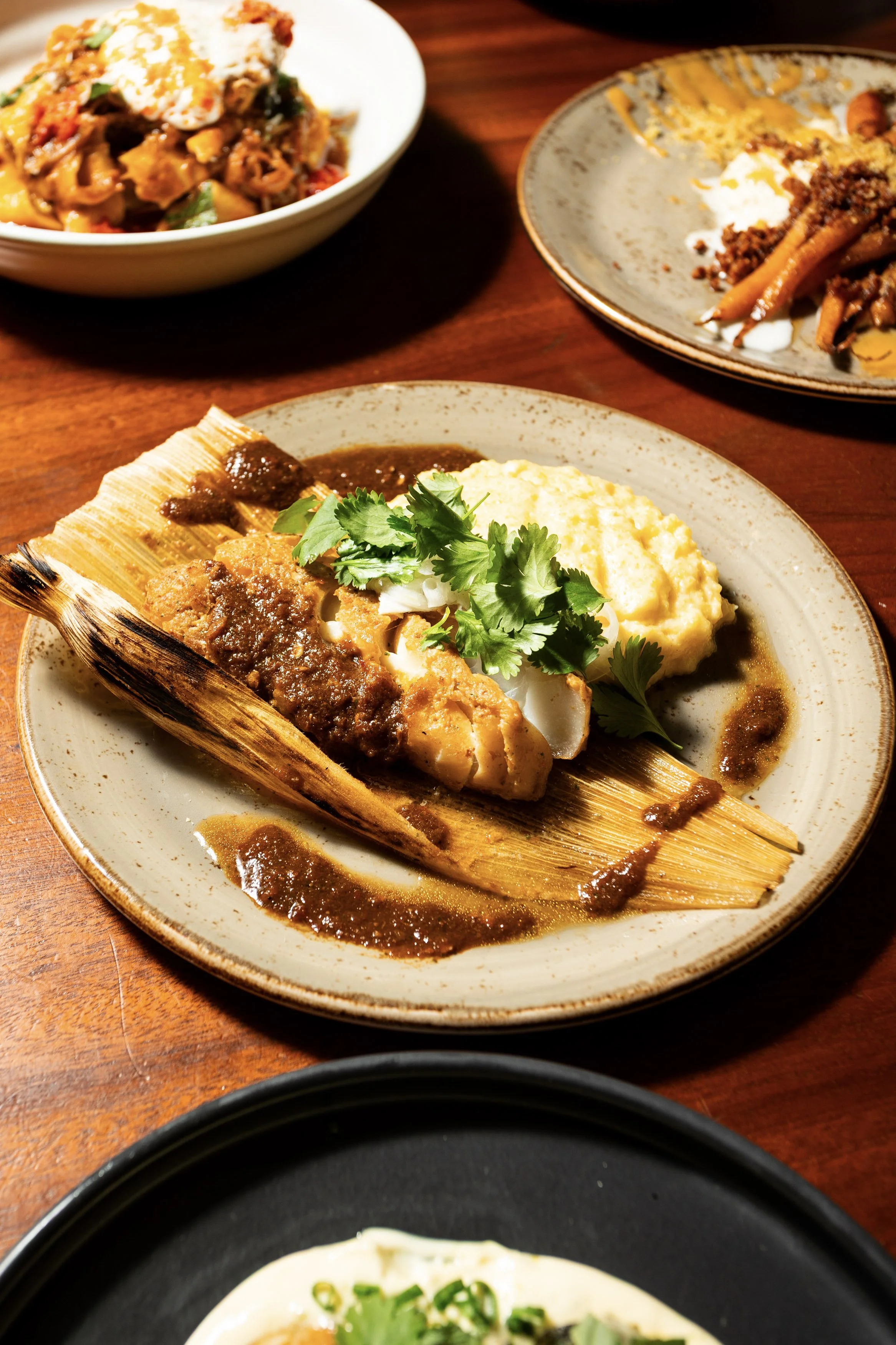 Plate with tamal topped with sauce, mashed potatoes, and cilantro, surrounded by three other dishes in bowls and plates on a wooden table.