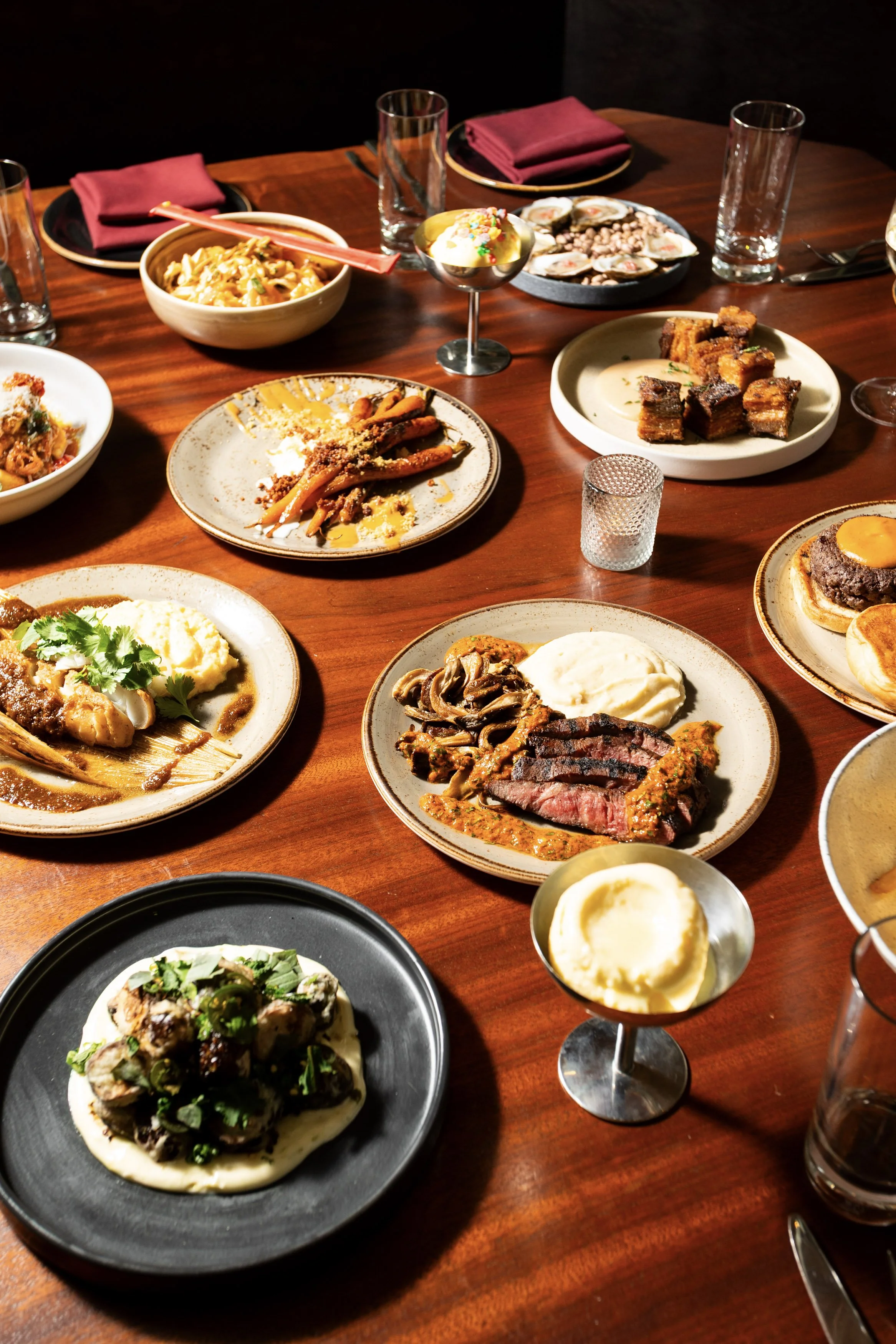 A wooden table filled with various plates of food, including mashed potatoes, steak, pasta, ribs, and salads, in a dimly lit restaurant setting.