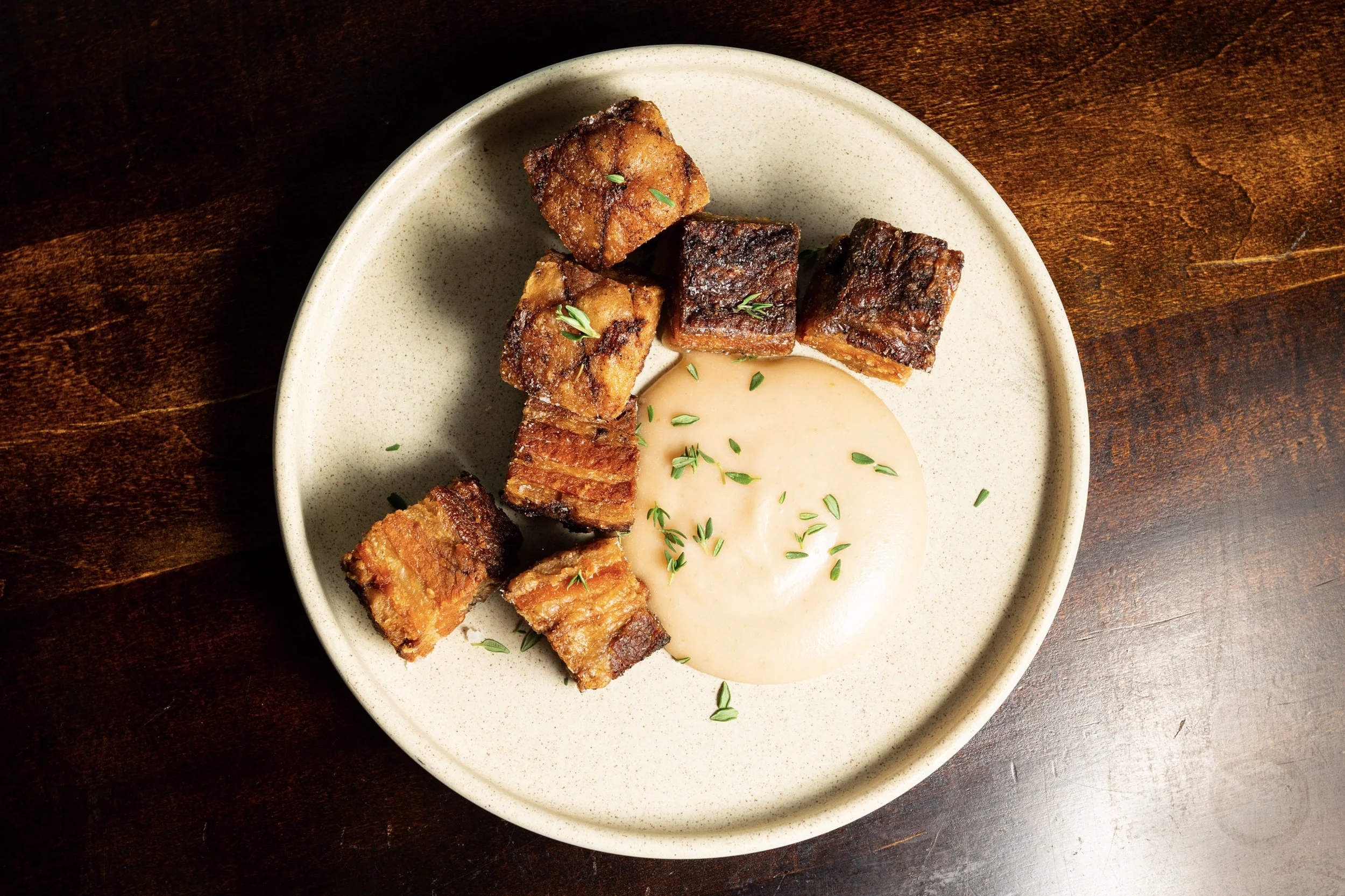 A beige bowl contains several pieces of crispy pork belly with garnish, served with a creamy sauce topped with chopped herbs, on a dark wooden surface.