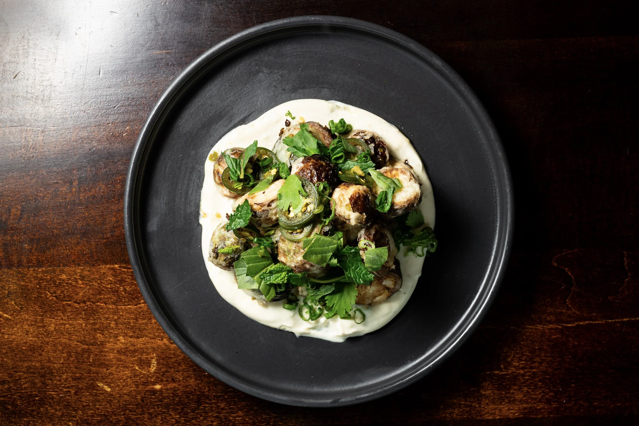 Plate of grilled chicken pieces topped with chopped green herbs and sliced jalapeños, served on a bed of white sauce on a black dish.