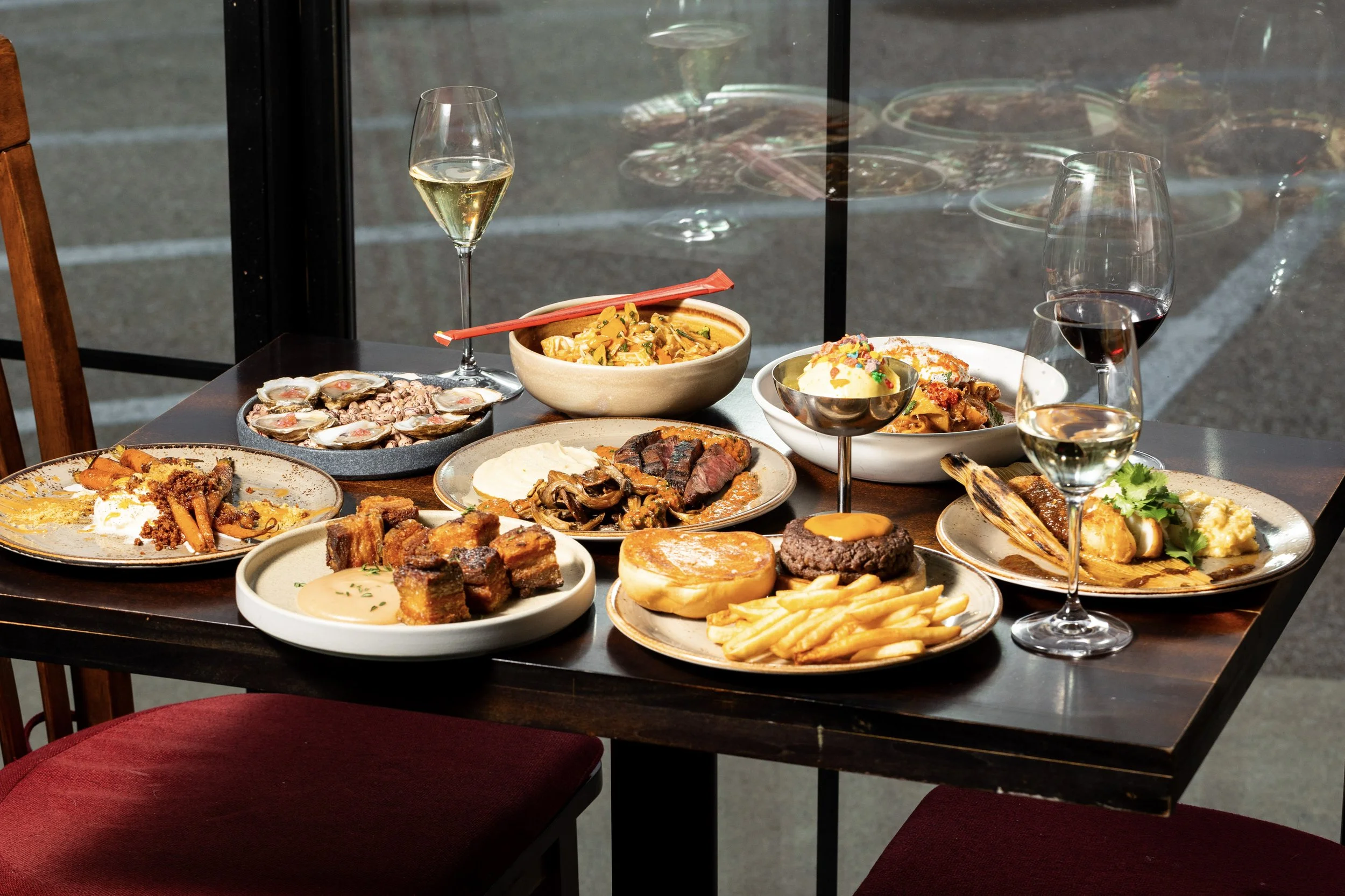 Various dishes including steak, fries, burgers, seafood, and desserts on a dark wooden table with glasses of white and red wine, set near a window.