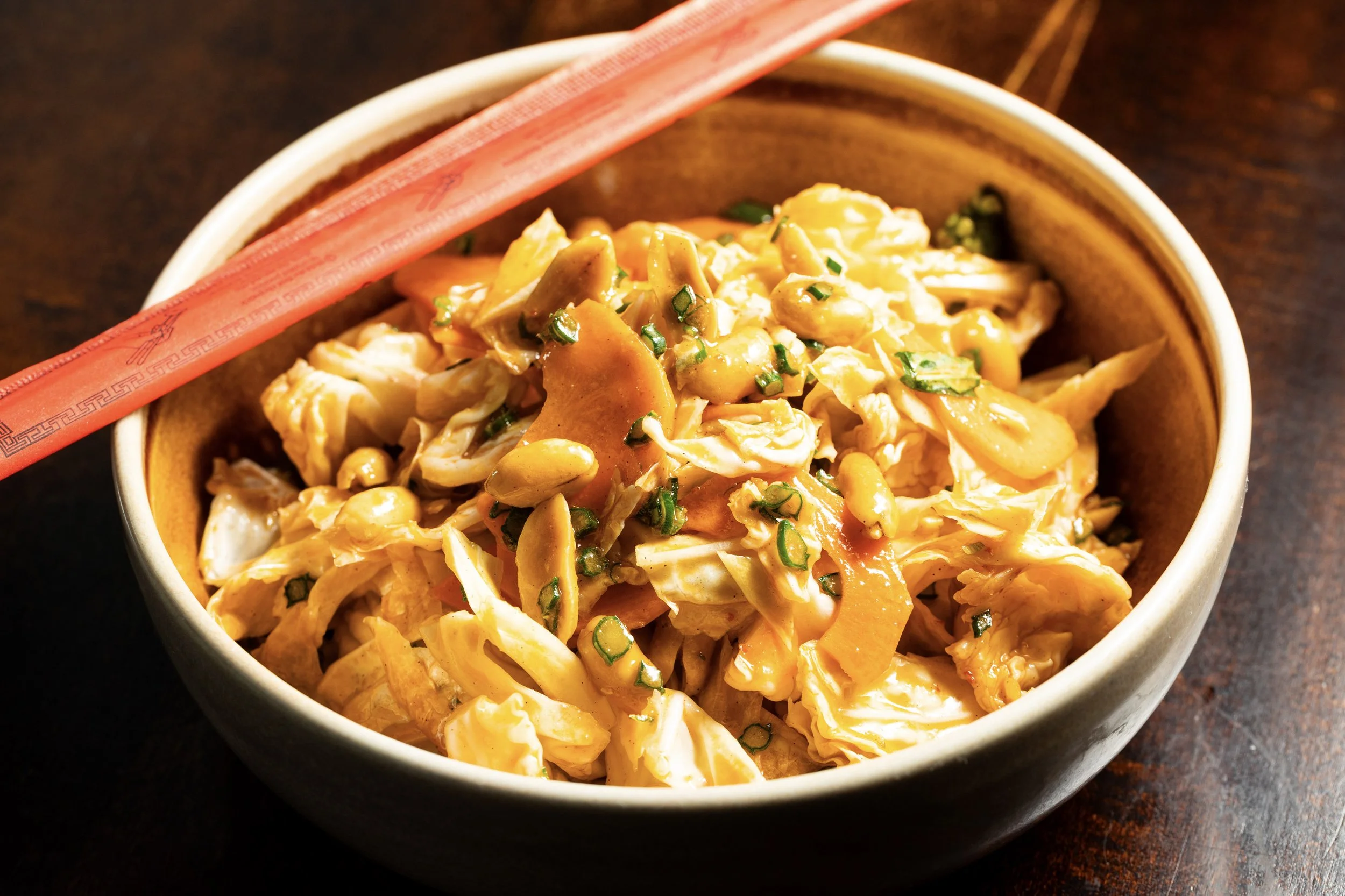 A bowl of Asian-style cabbage salad with chopped green onions and peanuts, with a pair of red chopsticks resting on the edge of the bowl.