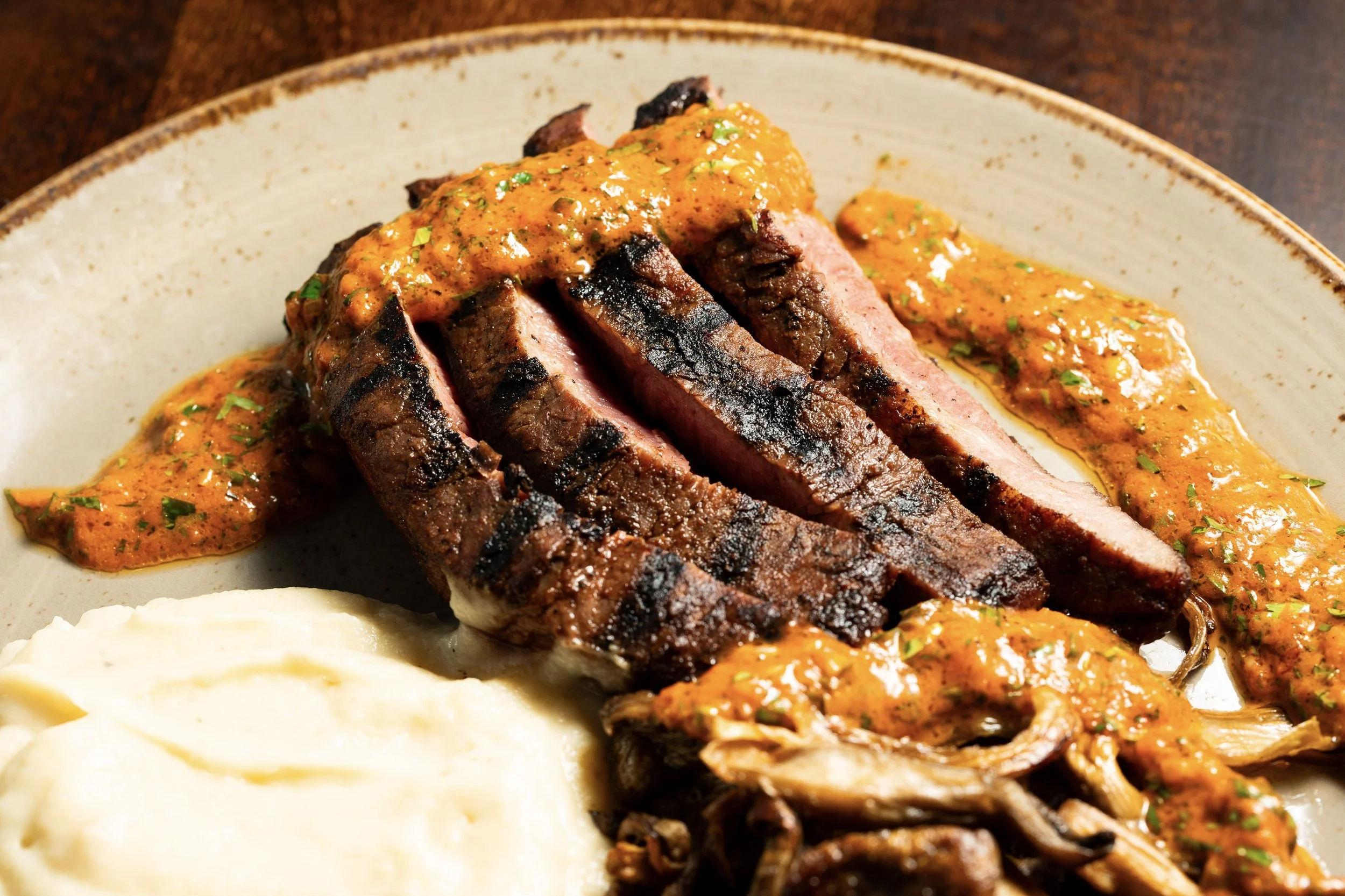 A plate of grilled steak with grill marks, topped with orange sauce, served with a side of mashed potatoes and sautéed mushrooms with sauce.