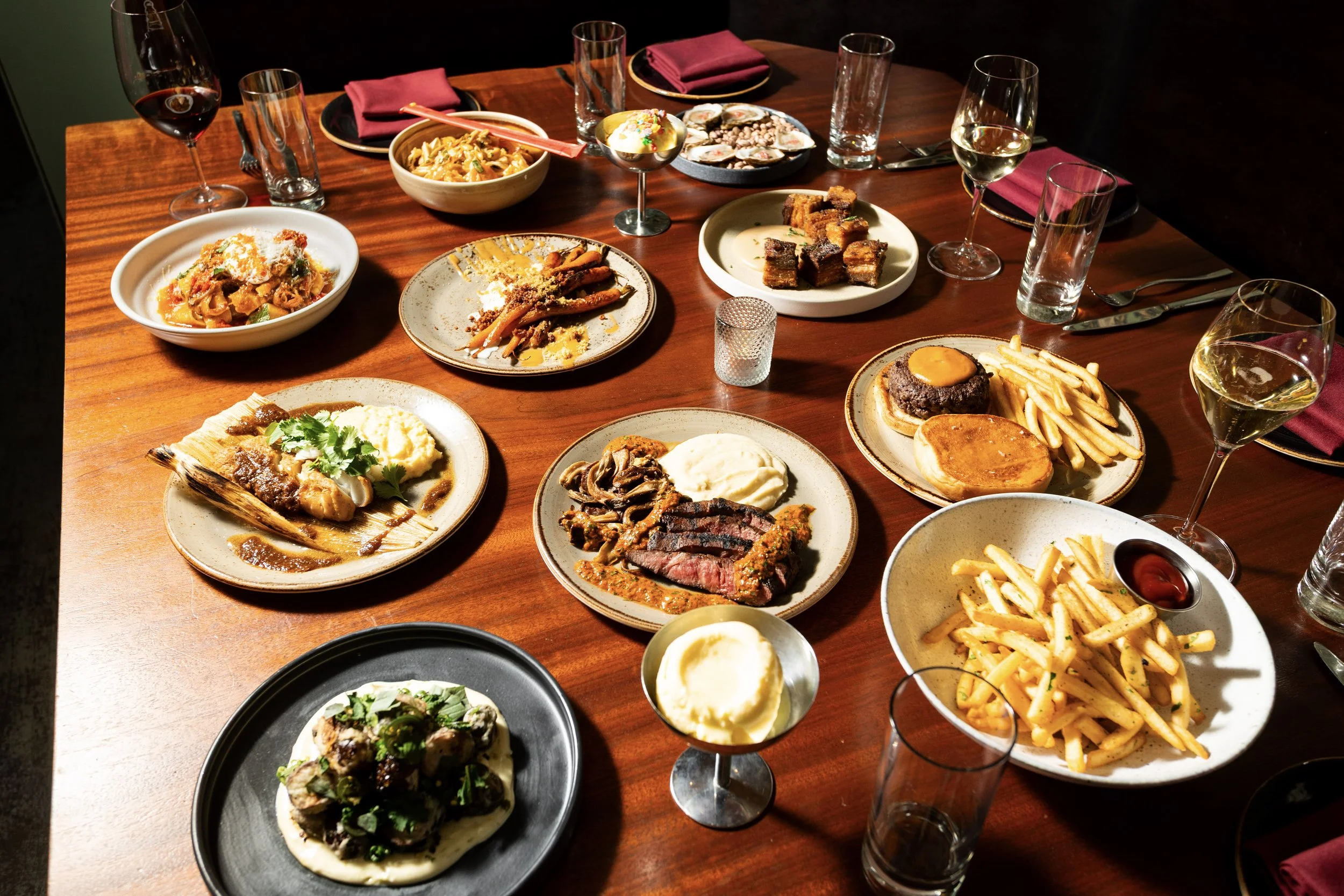 A dinner table filled with various plates of food, including pasta, steak with mashed potatoes, French fries, burger with an egg, and salads, along with glasses of wine and water, set for multiple people.