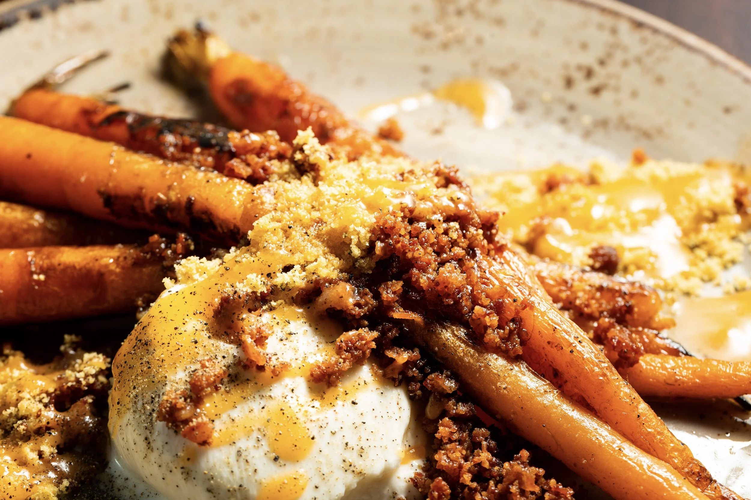 Close-up of roasted carrots topped with ground meat, breadcrumbs, and melted cheese, with a poached egg and black pepper.