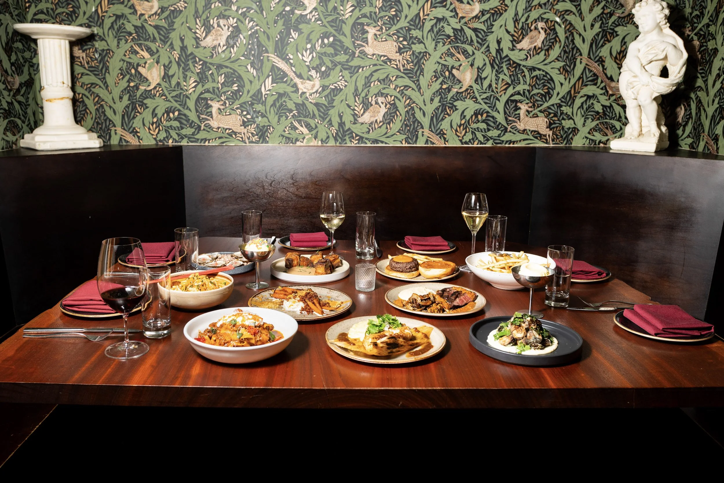 A dinner table set with various plates of food, glasses of wine, and utensils, in a restaurant with dark wood accents and green patterned wallpaper with animals and plants.