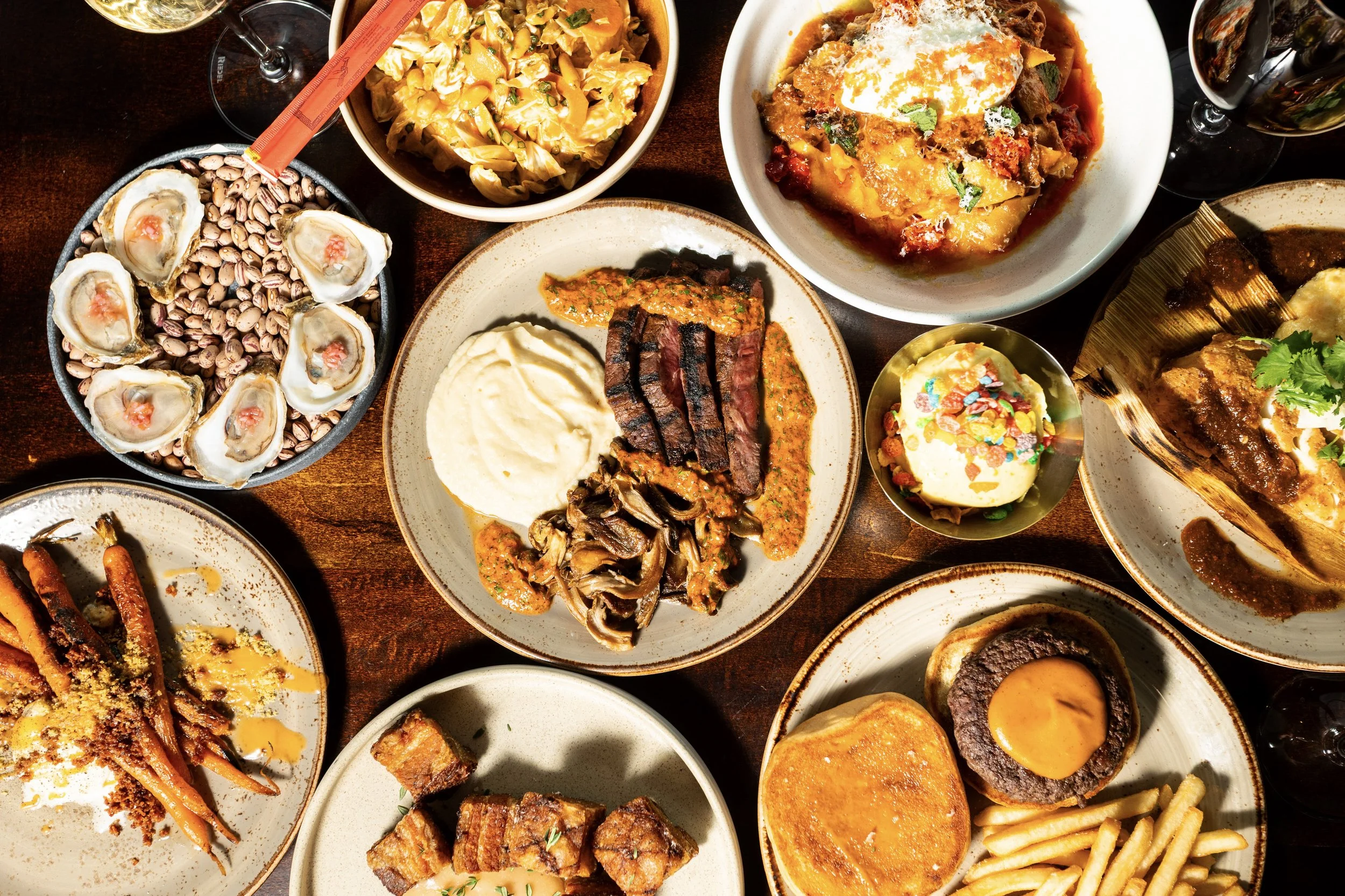 A variety of dishes including oysters, pasta, steak with sauce, curry, a baked dish, meatloaf, fried carrots, and a dessert, along with glasses of red wine on a wooden table.