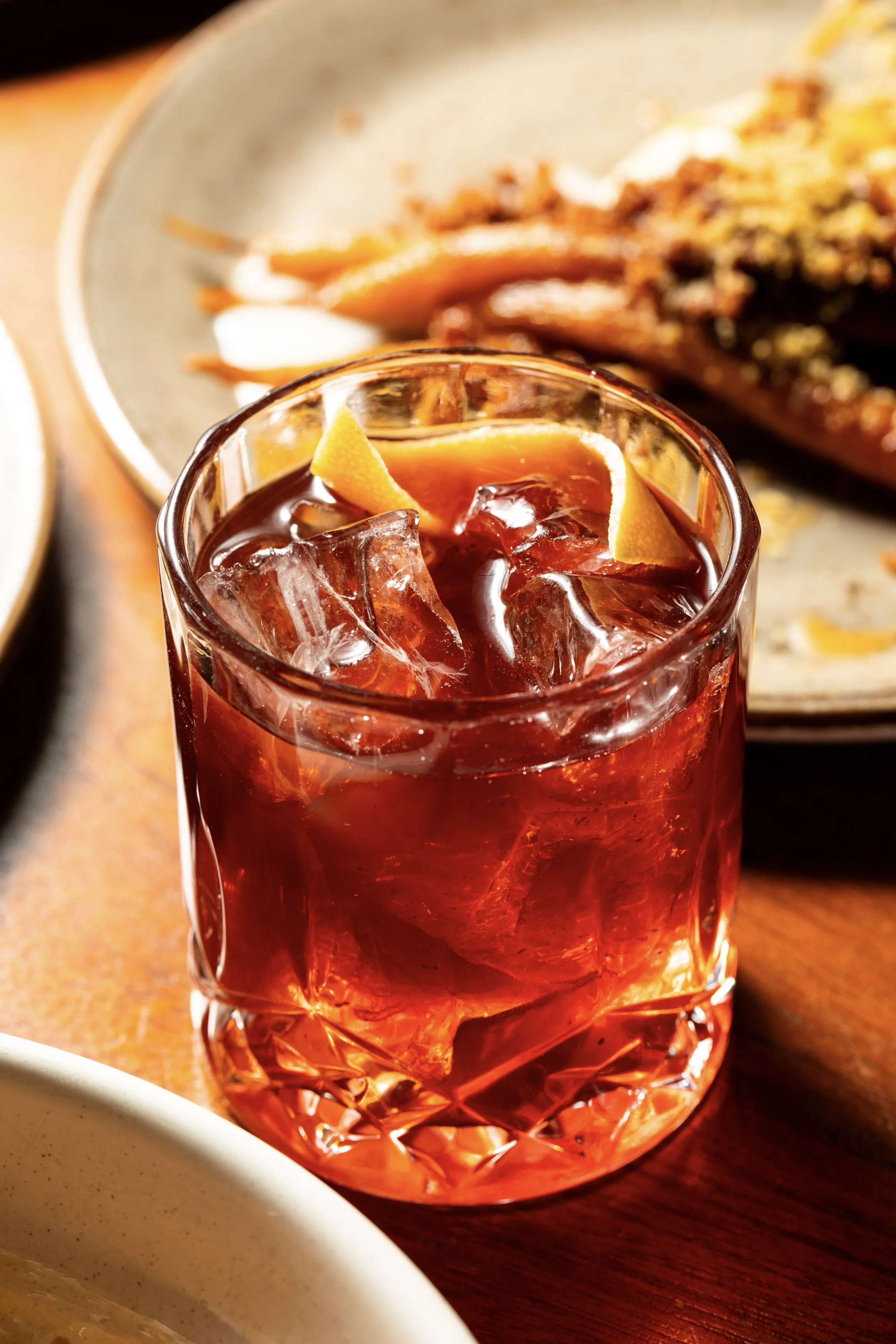 A glass of iced sweet tea with lemon wedges on top, on a wooden table, with a plate of food in the background.