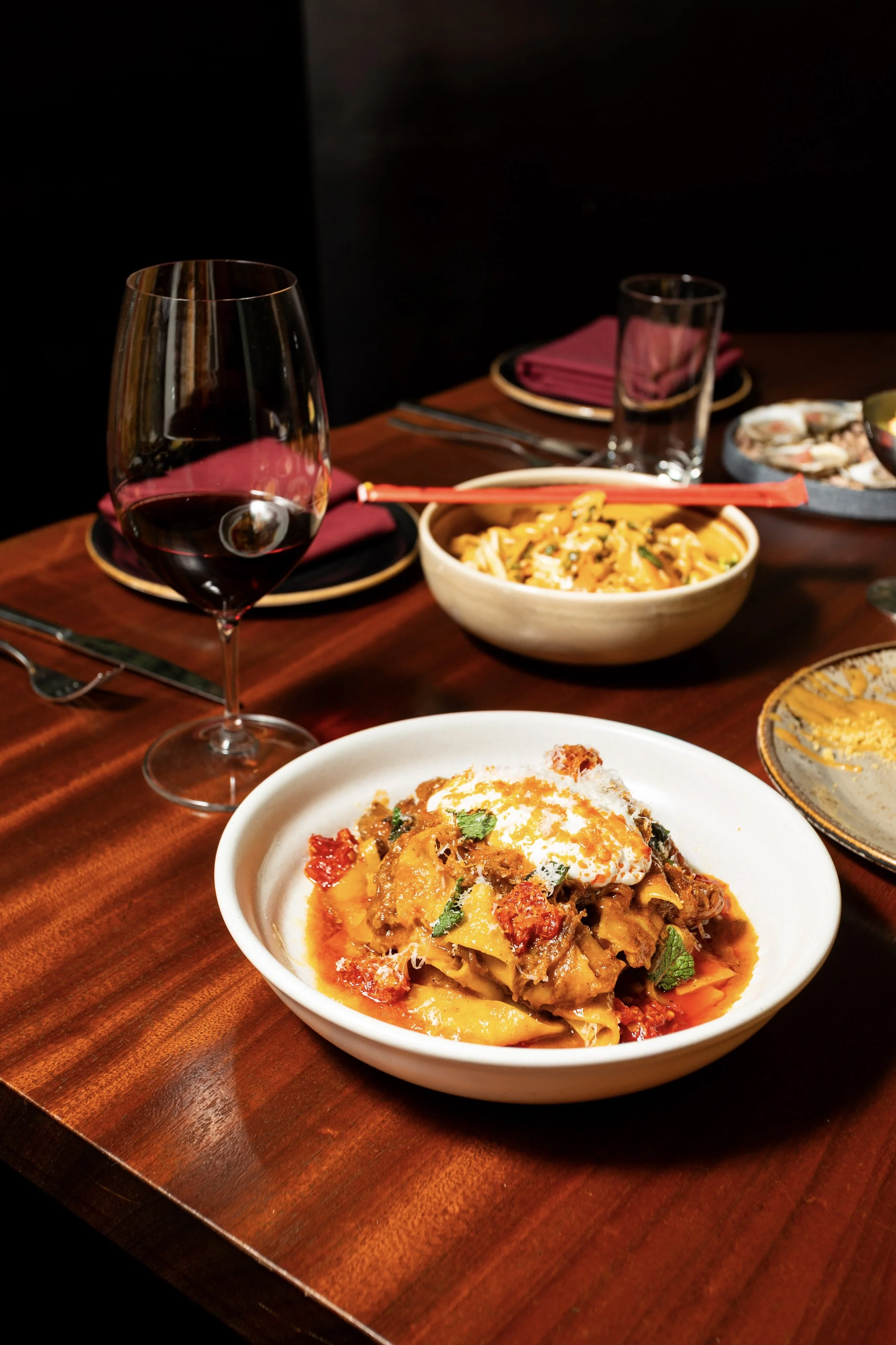 A dinner table with a glass of red wine, a bowl of pasta, and a bowl of stew topped with an egg, set on a wooden table with plates, silverware, and napkins.