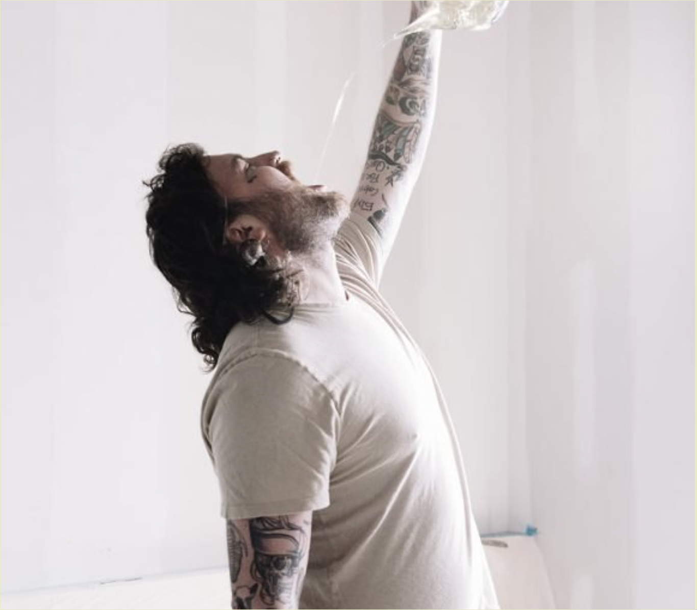 Man with tattoos and long dark hair, wearing a light-colored T-shirt, tilting his head back and pouring a clear liquid from a plastic bag into his mouth.