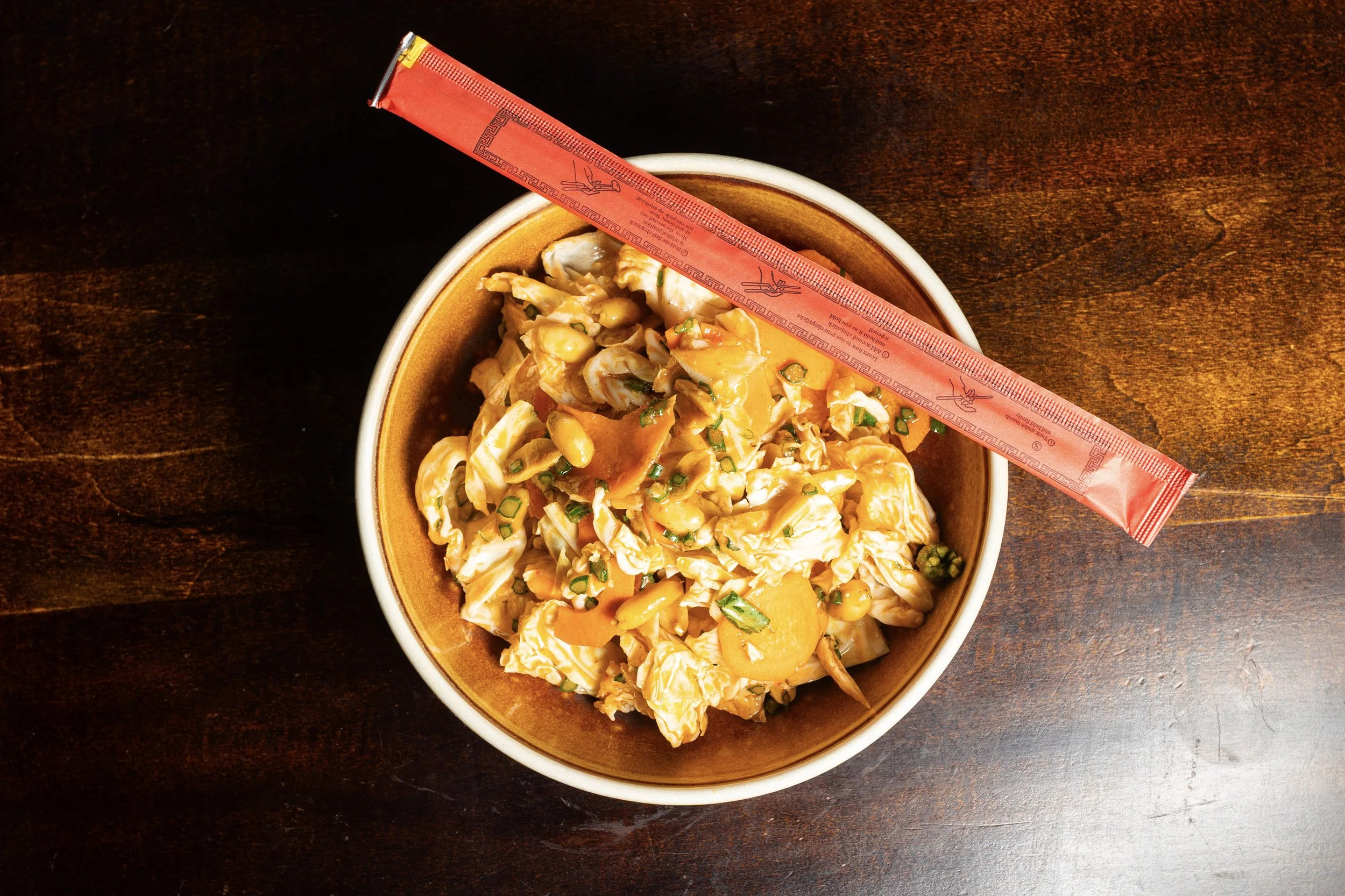 A bowl of Asian-style chopped cabbage salad with scallions and peanuts on a dark wooden table, with a red chopstick rest and chopsticks resting on the bowl.