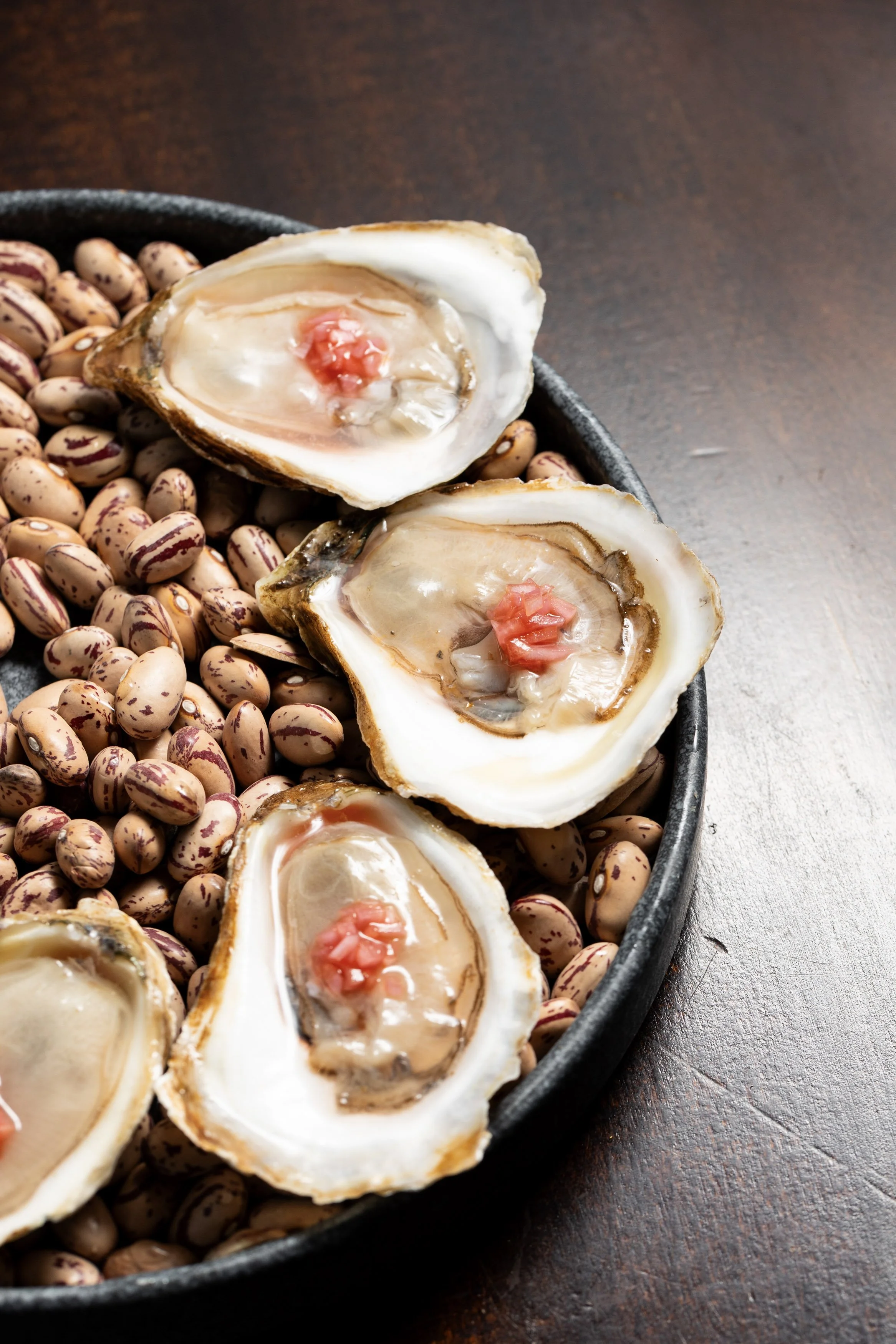Fresh oysters served on a black plate with pinto beans, placed on a dark wooden surface.