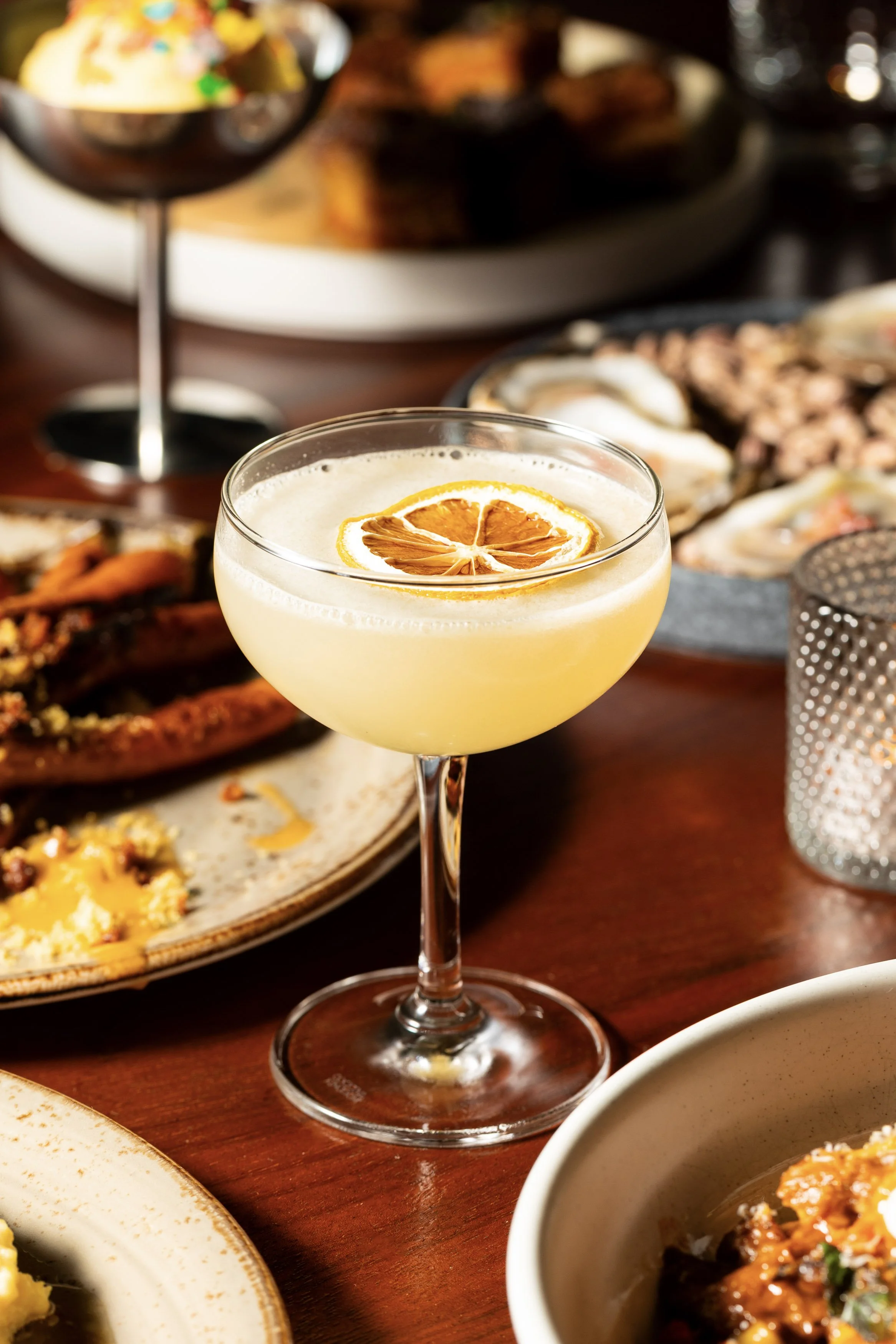 A cocktail in a coupe glass with a slice of dried orange on top, placed on a wooden table with various dishes in the background, including seafood and appetizers.