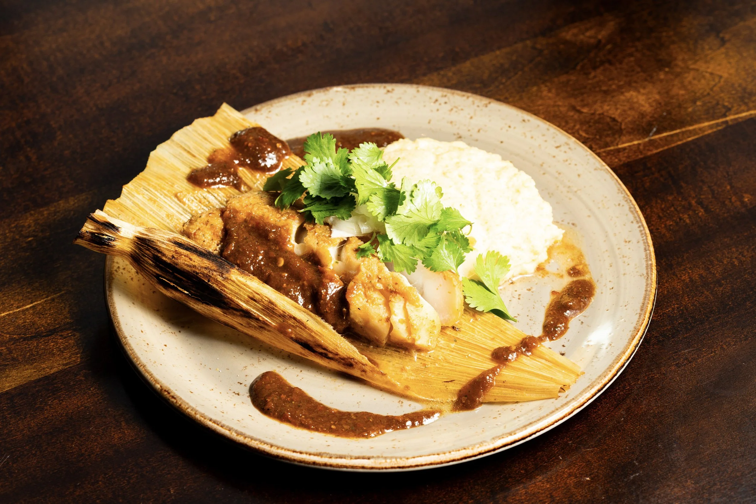 A plate of Mexican dish featuring tamale topped with meat sauce, garnished with cilantro, served alongside mashed potatoes or similar, on a rustic wooden table.