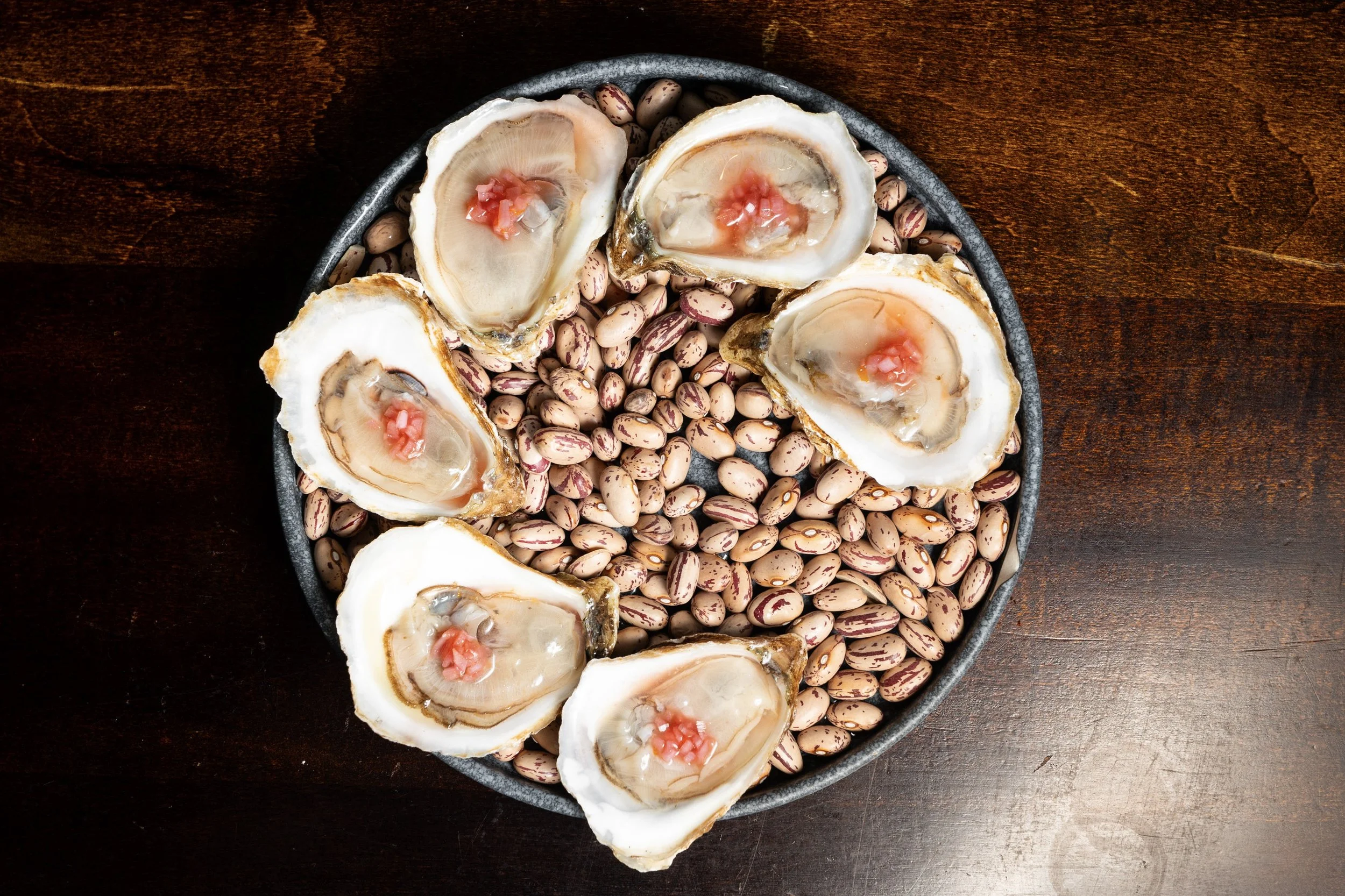 A round black plate filled with cooked pinto beans and topped with five raw oyster shells