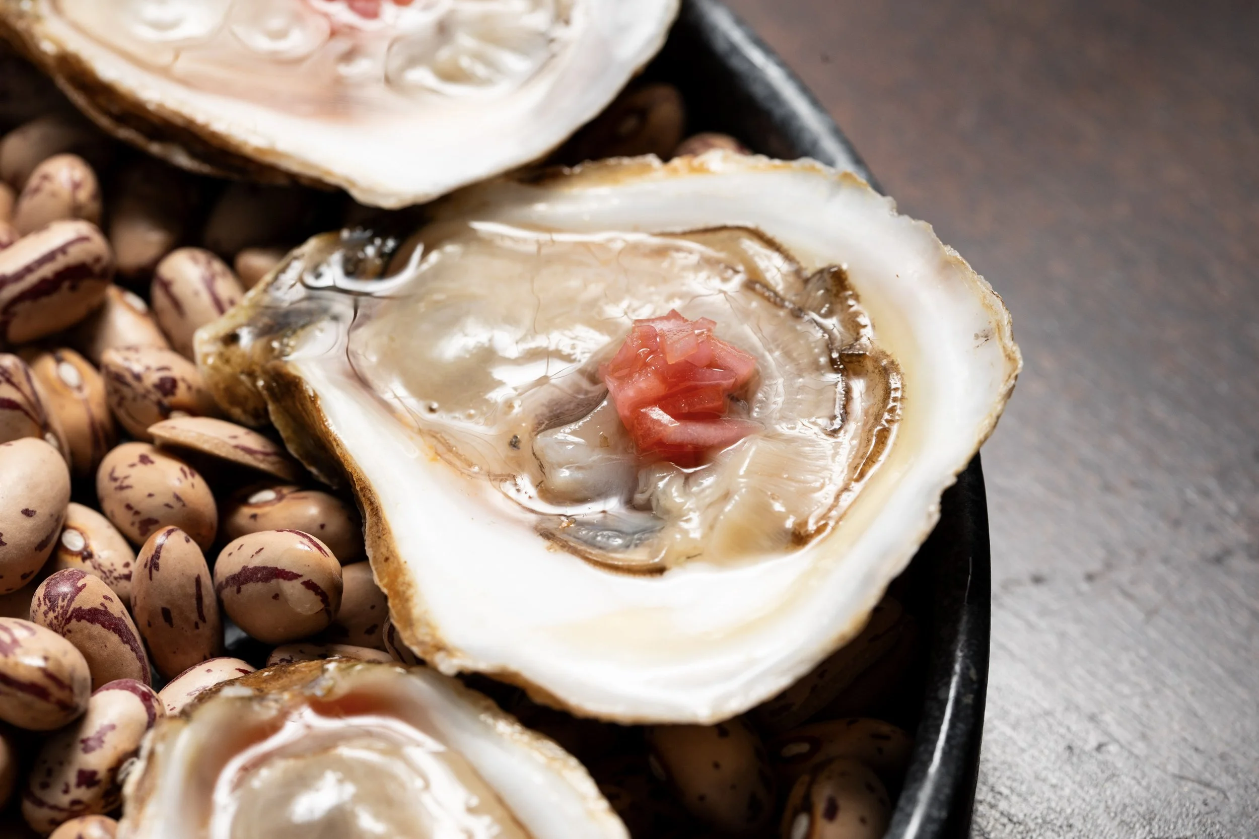 Close-up of a fresh oyster on the half shell with a small piece of pink tomato, served on a bed of pinto beans in a black dish.