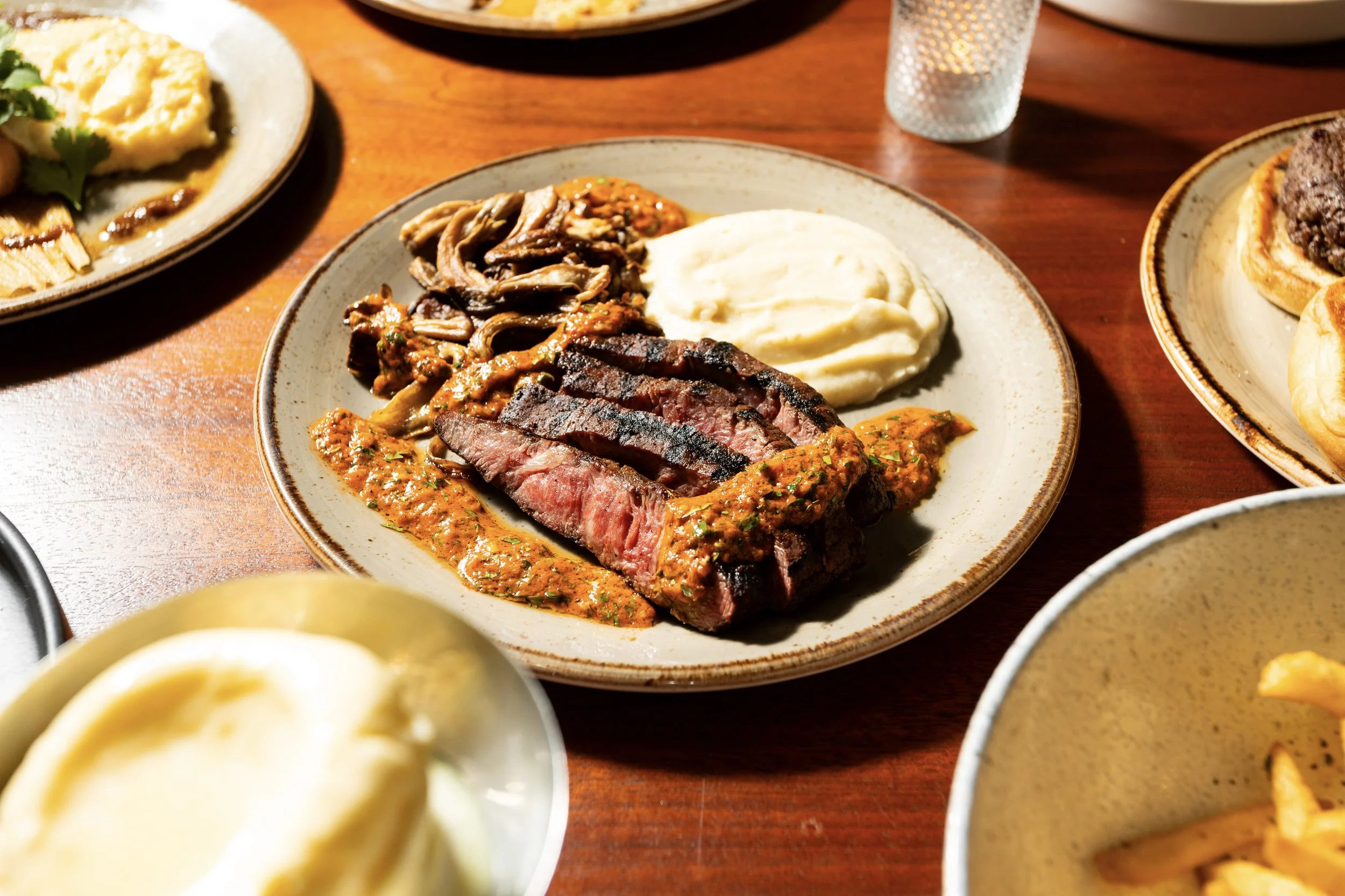 Plate with sliced grilled steak, mashed potatoes, sautéed mushrooms, and a drizzle of sauce on the steak, on a wooden table with other plates of food.