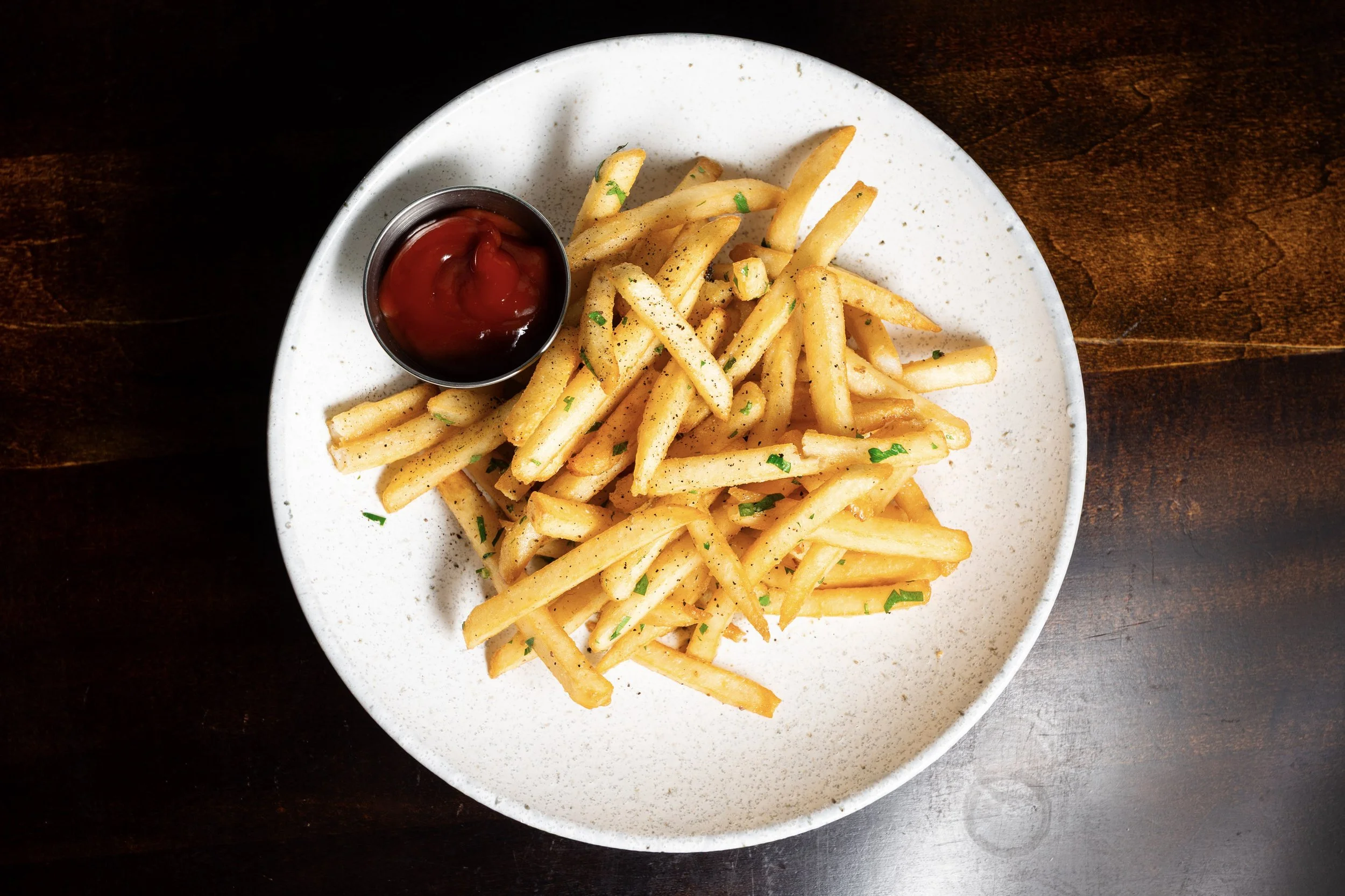 A white plate with seasoned French fries garnished with chopped parsley and a small metal cup of ketchup, set on a dark wooden table.