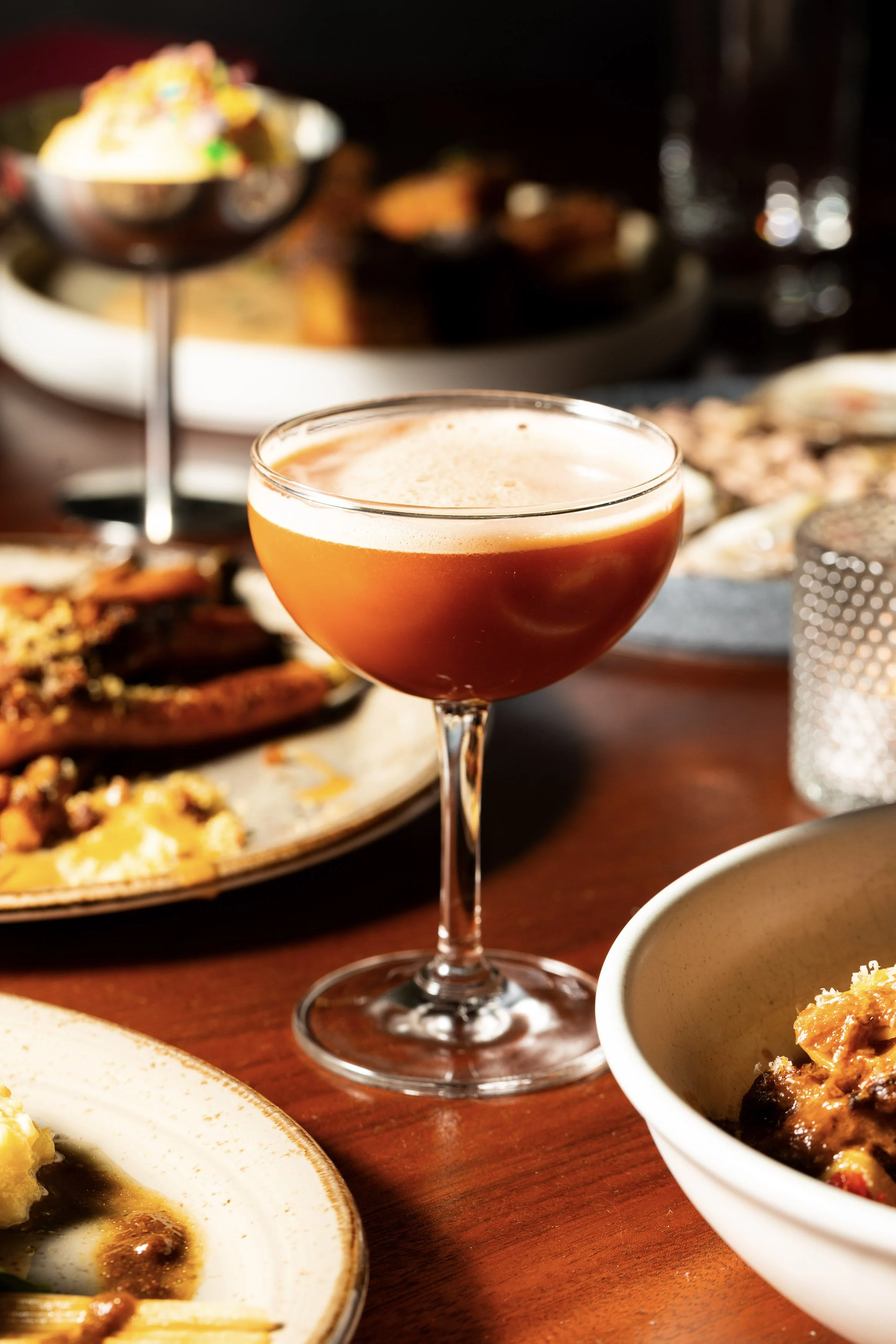 A cocktail glass filled with a frothy orange-colored drink on a wooden table among plates of food and a small glass candle holder.