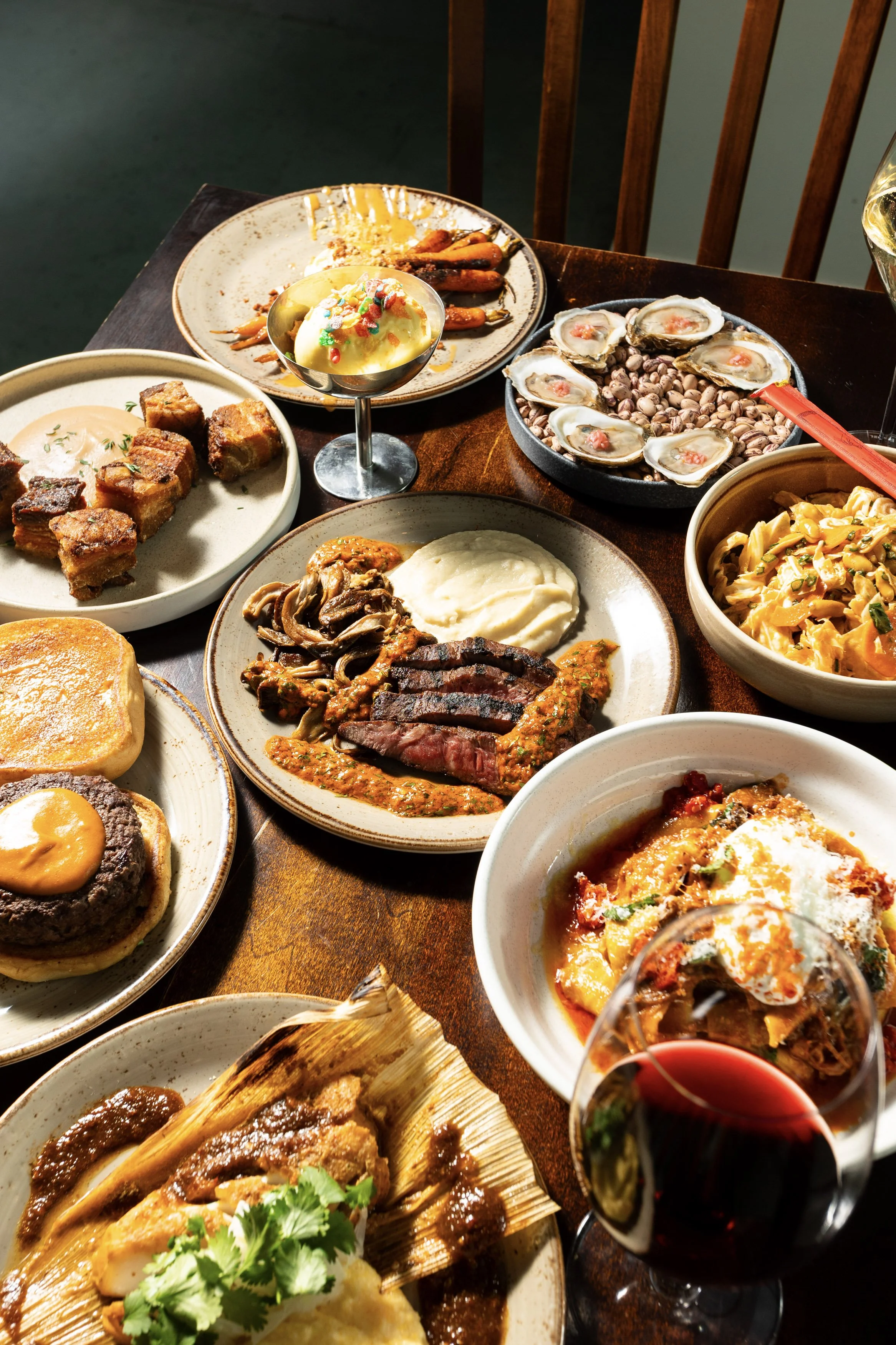 A variety of dishes on a wooden table, including grilled meats, pasta, seafood, and a glass of red wine.