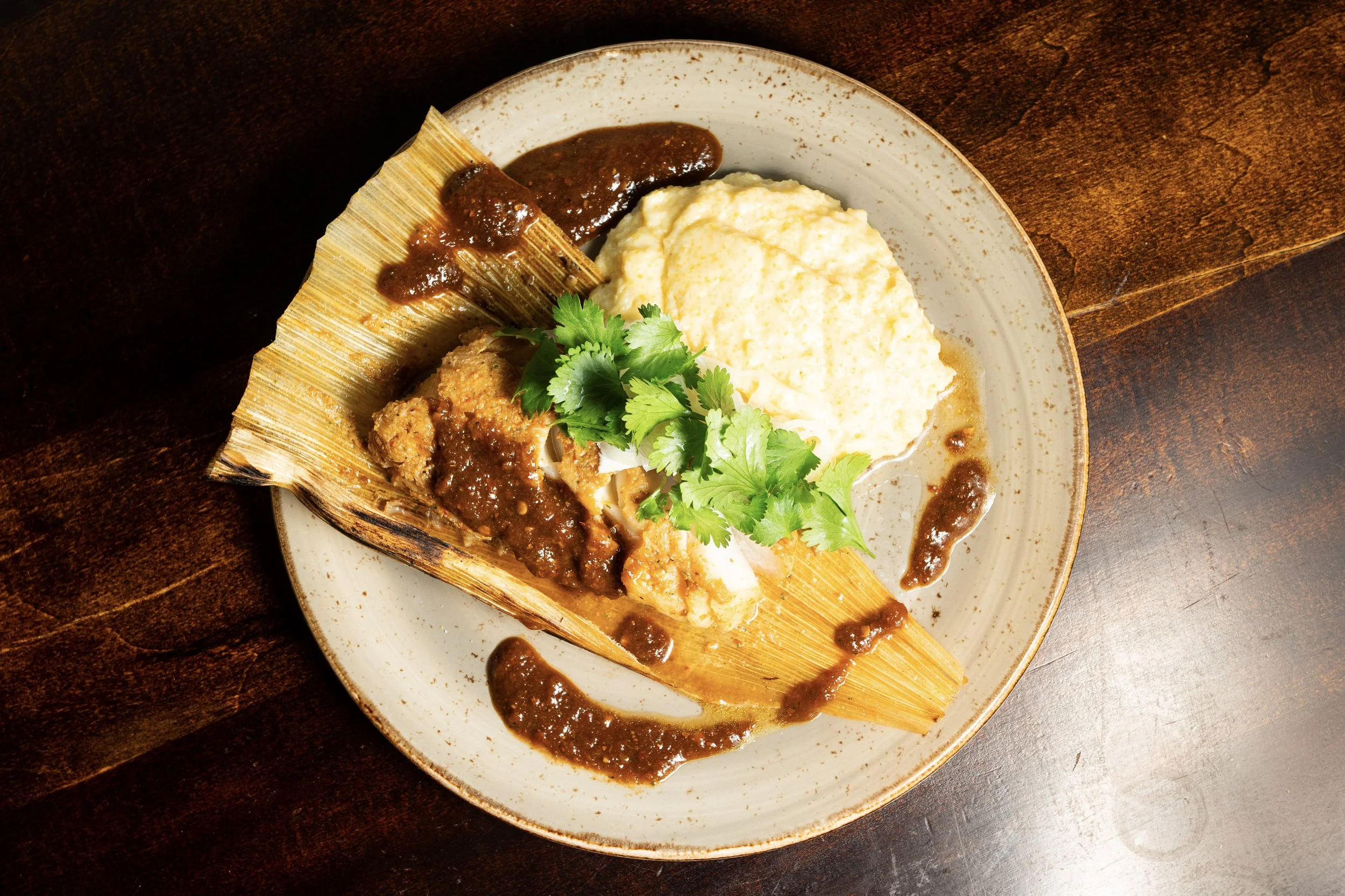 Plate of Mexican dish with tamale covered in sauce, served with mashed potatoes or cheese, garnished with fresh cilantro on a ceramic plate on a dark wooden surface.