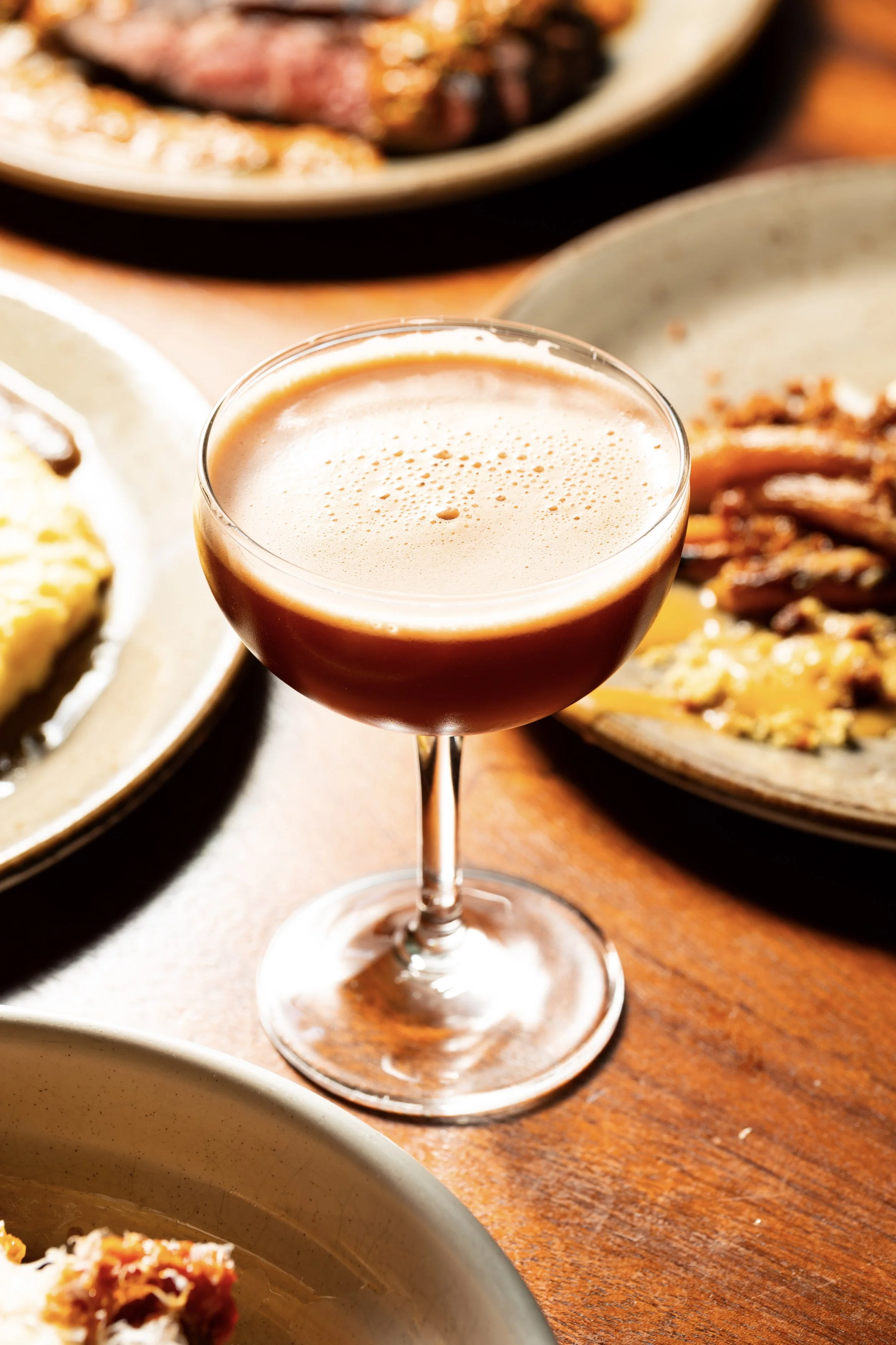 A cocktail drink in a coupe glass on a wooden table surrounded by plates of food.