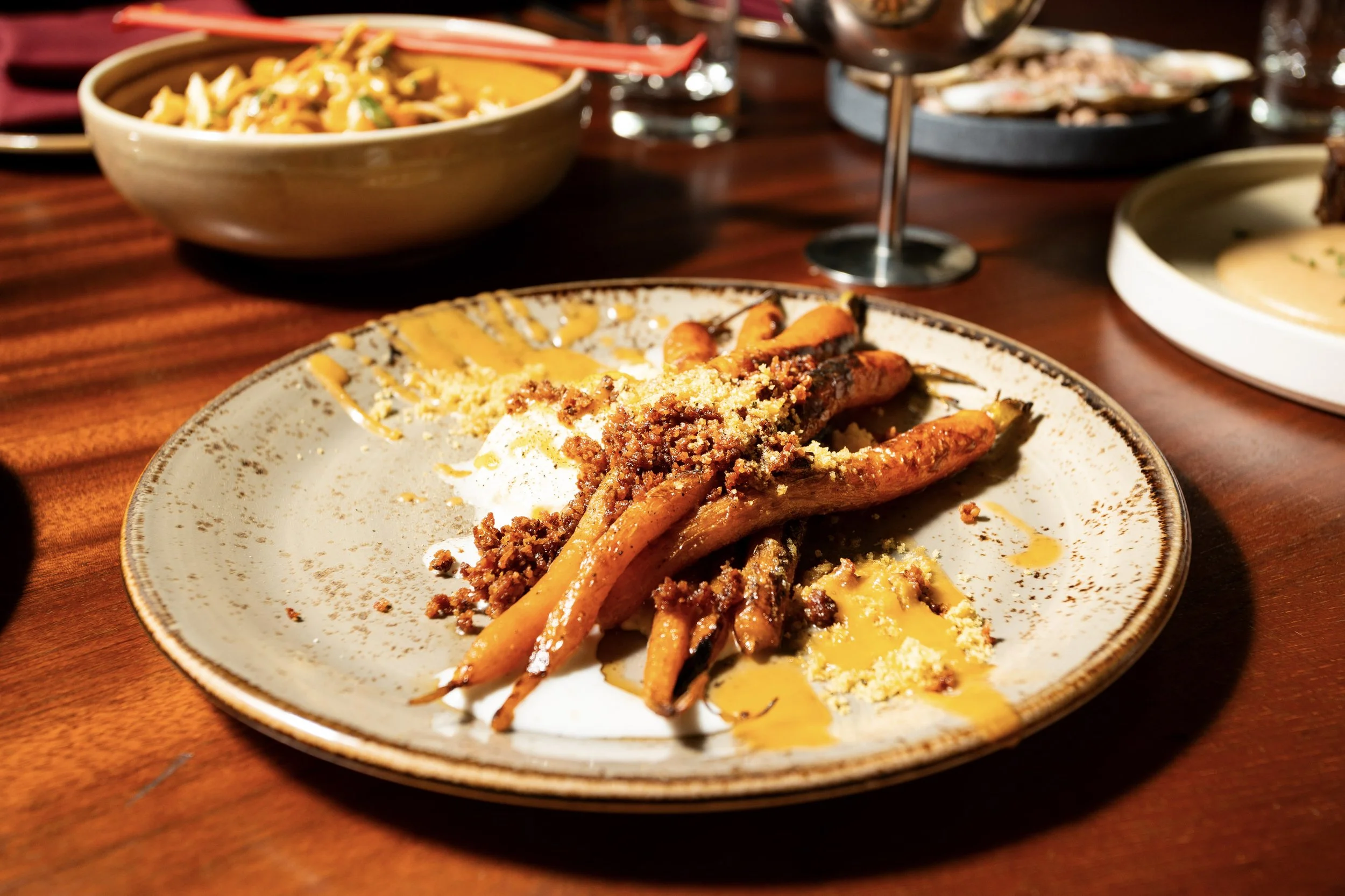 A plate with roasted carrots topped with ground meat and crumbled cheese, served with sauce and garnished with herbs, on a wooden table surrounded by bowls and glasses.