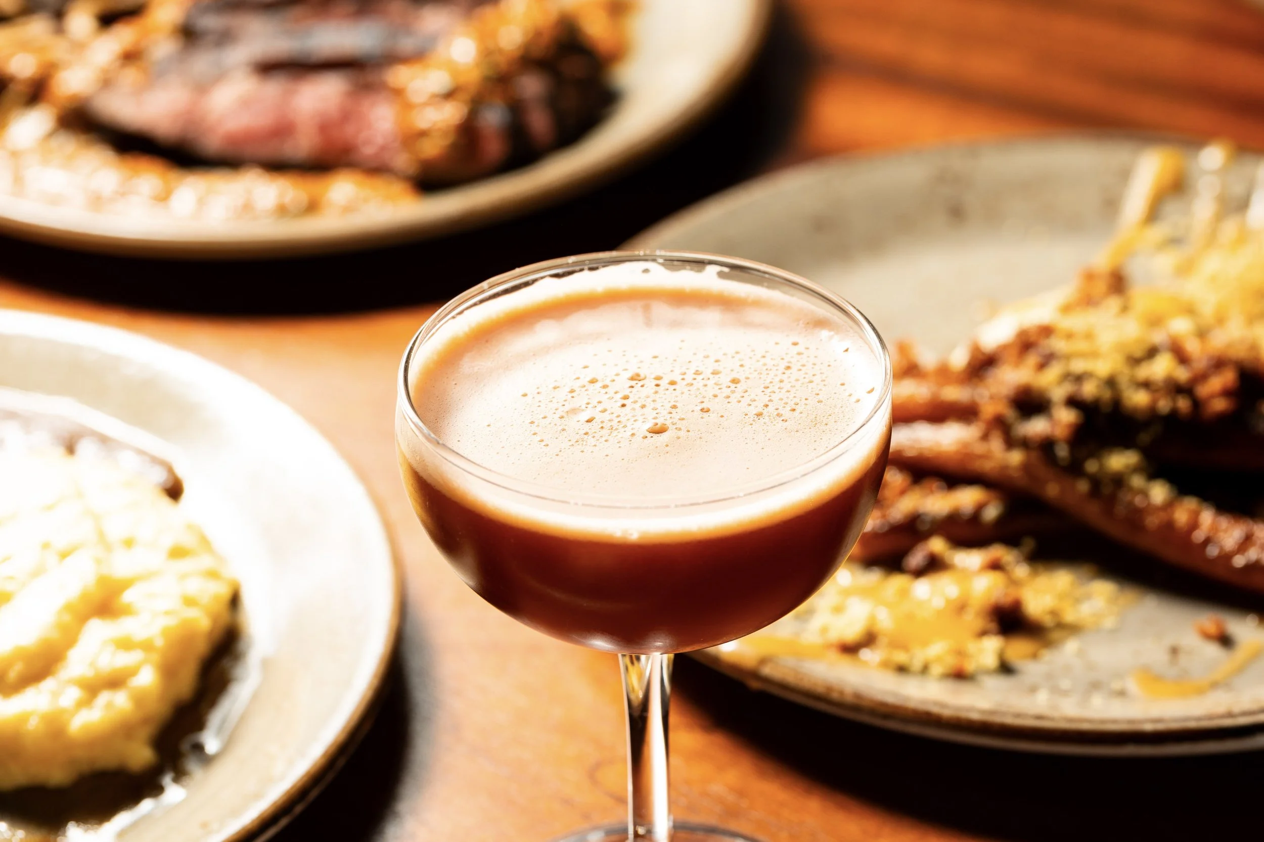 A cocktail glass filled with a creamy, frothy beverage surrounded by plates of food, including steak and eggs, on a wooden table.