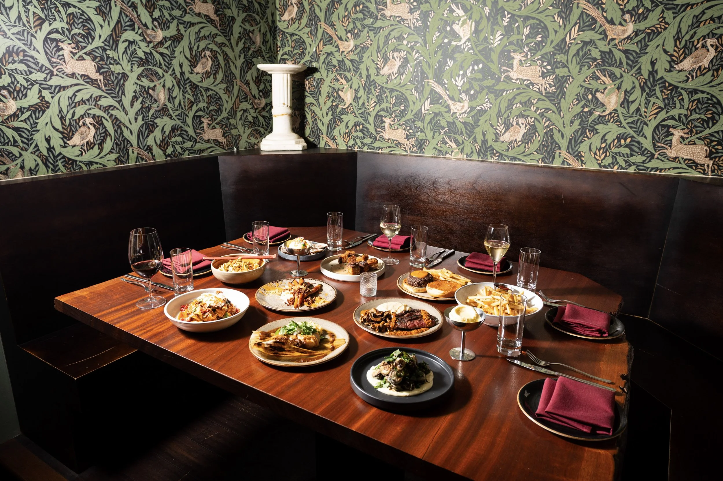 A wooden dining table set for a meal with various plates of food, wine glasses, and napkins, in a room with tropical leaf and bird patterned wallpaper and a white pedestal on the corner wall.