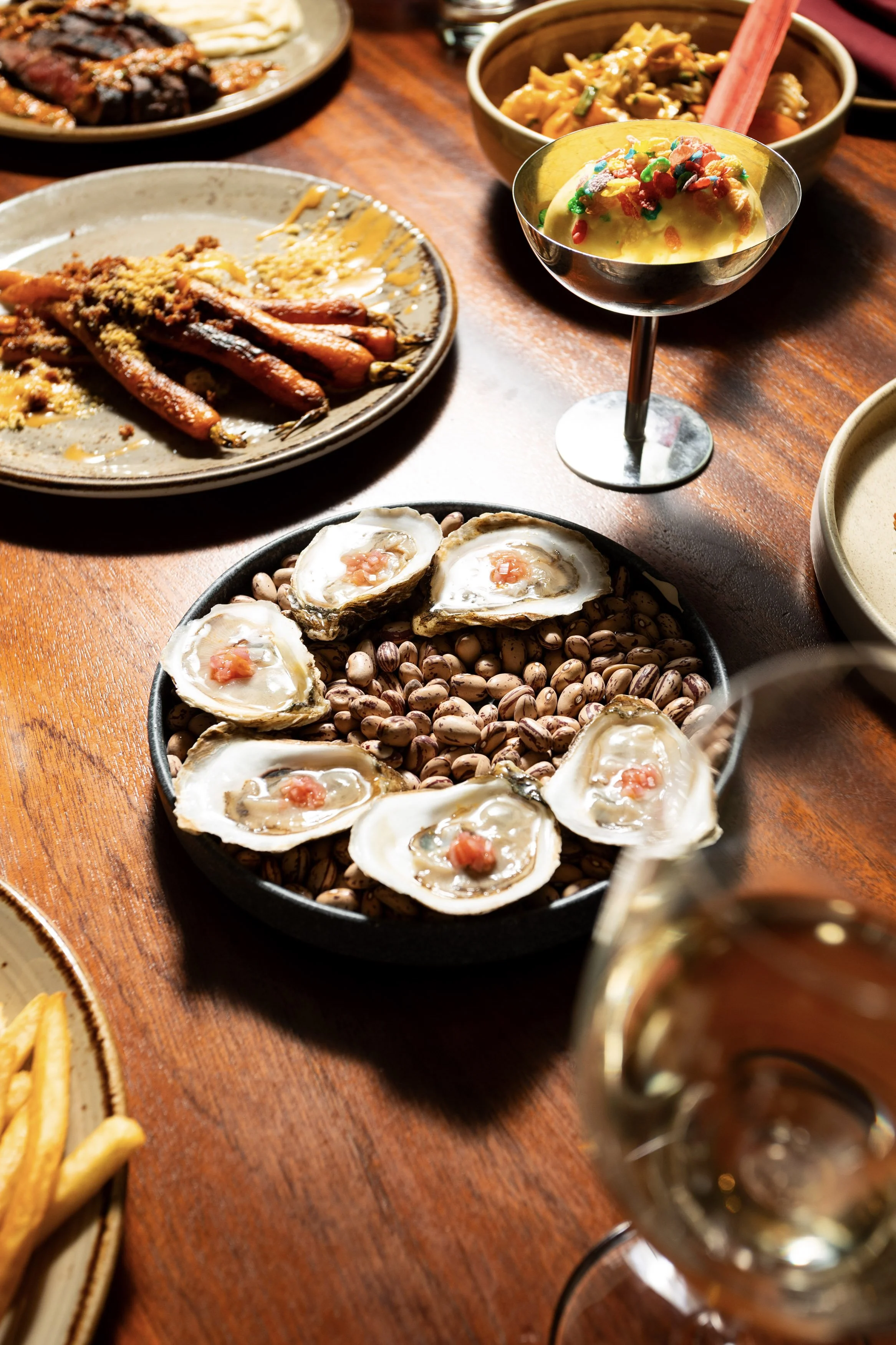 A variety of seafood dishes on a wooden table, including oysters on a bed of beans, grilled chicken wings, and a dessert topped with colorful sprinkles, with a glass of white wine.