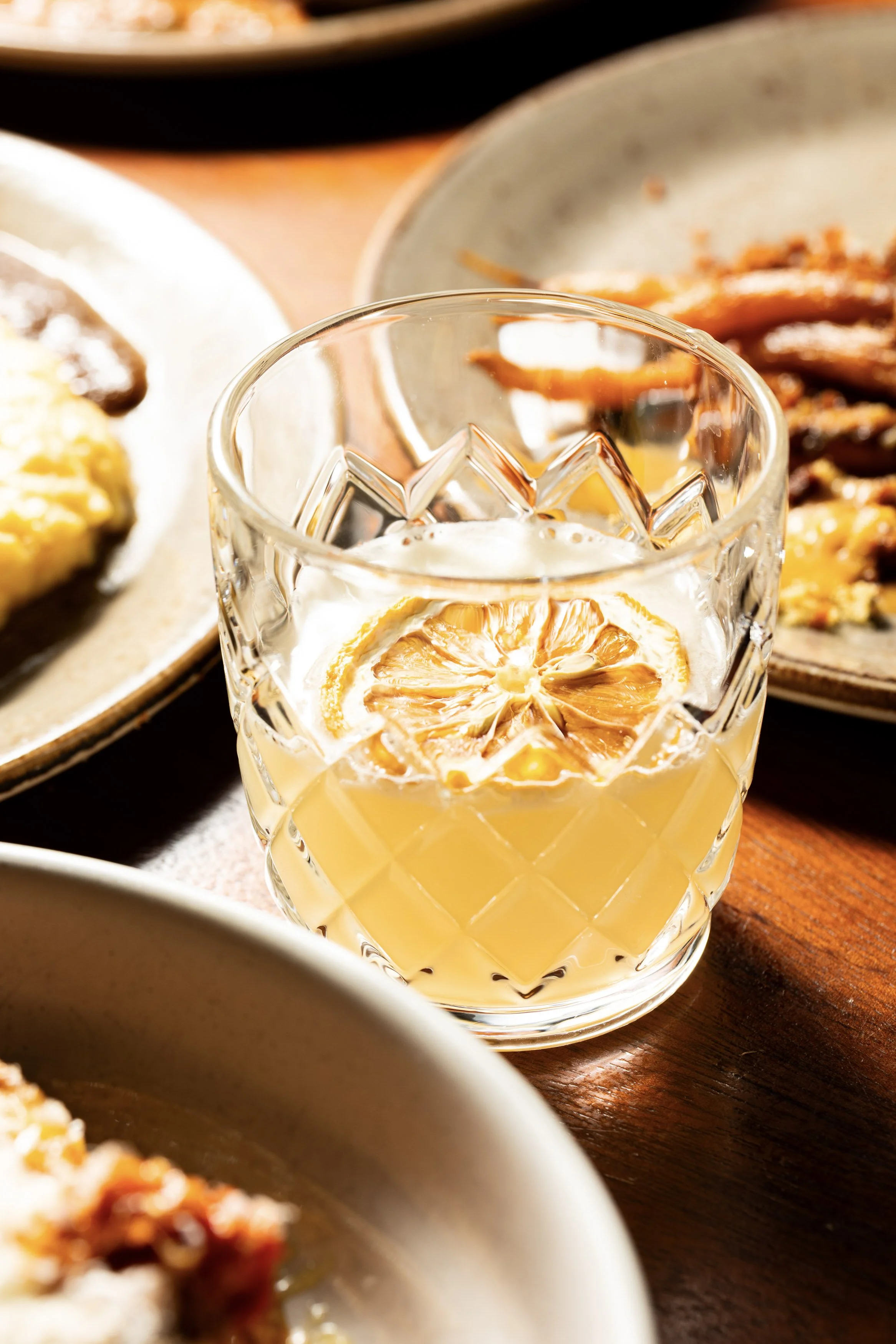 A glass of lemonade with a slice of lemon on top, placed on a wooden table with plates of food in the background.