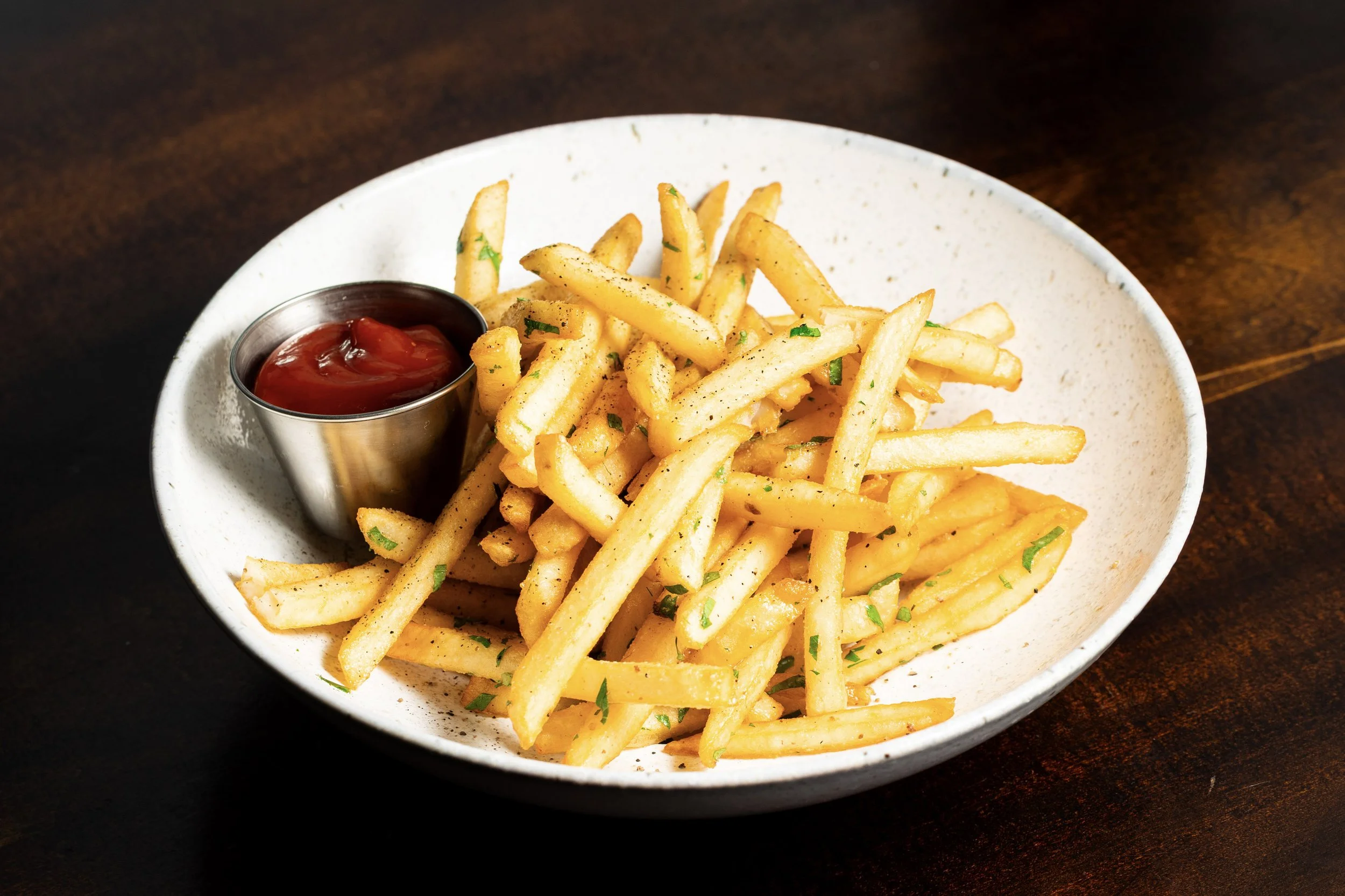 French fries with chopped herbs and a small cup of ketchup on a white plate.