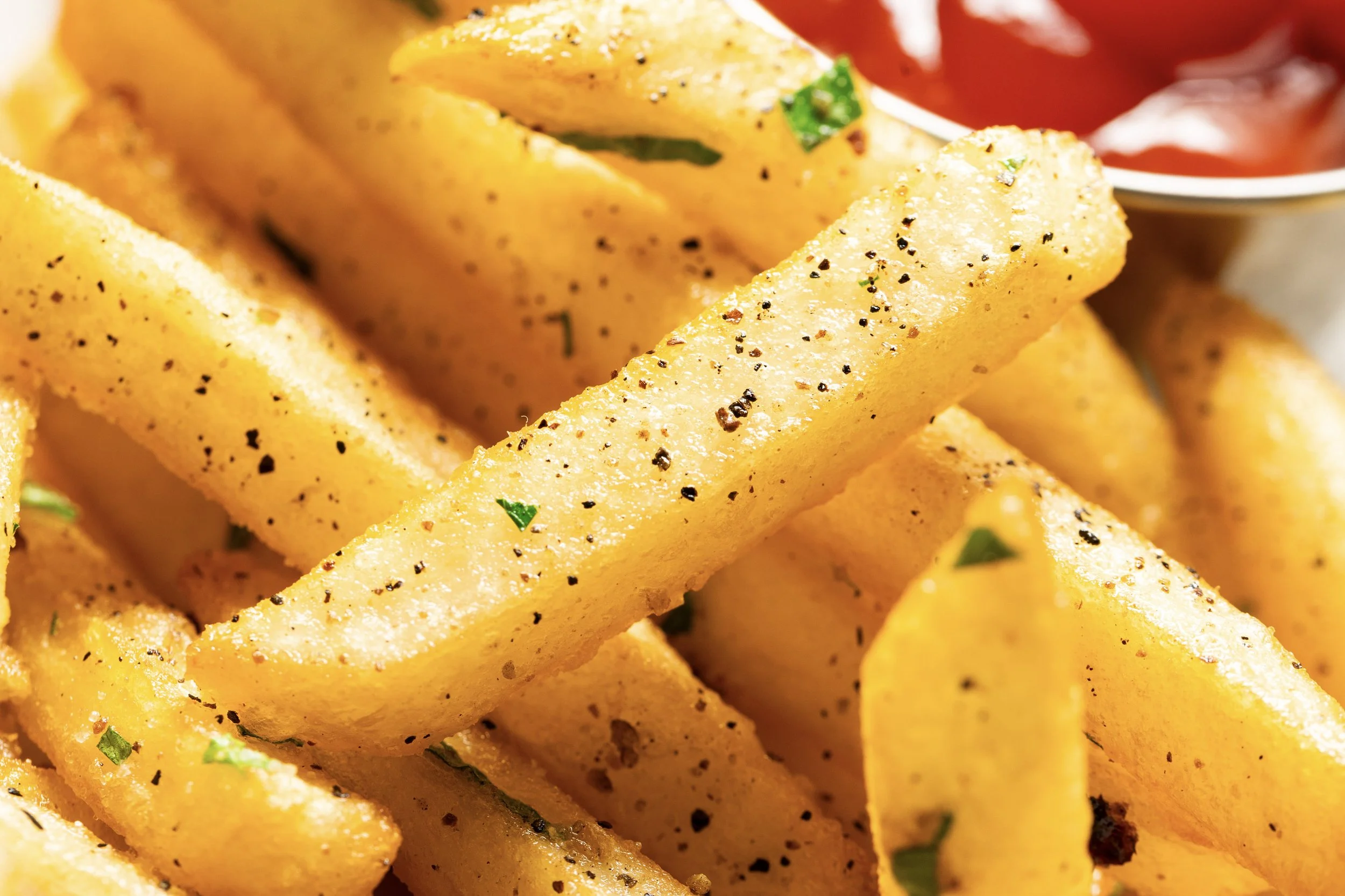 Close-up of crispy French fries sprinkled with black pepper and herbs, with a cup of ketchup in the background.