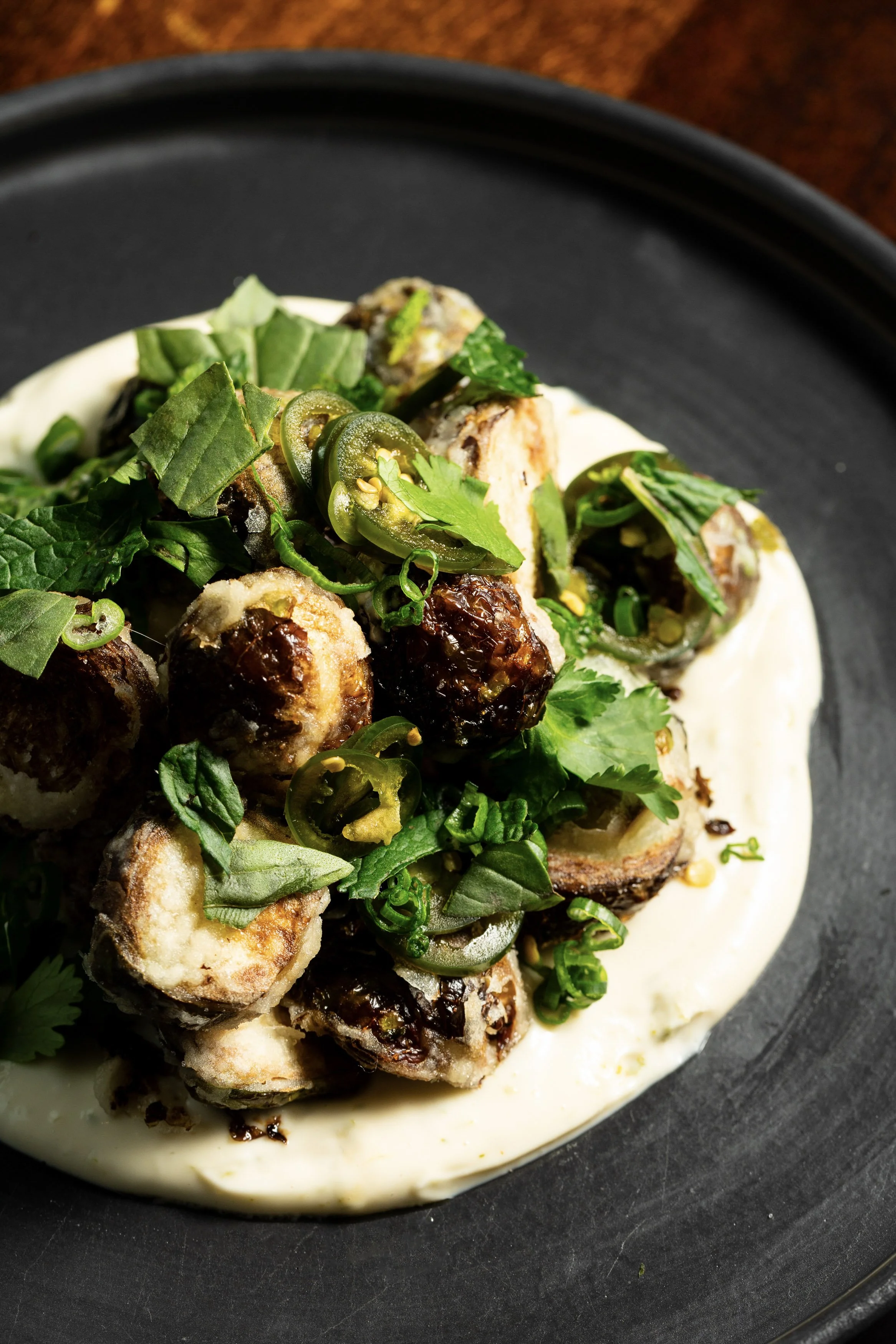 Close-up of a plate with fried pieces topped with sliced jalapeños, chopped green onions, and cilantro, served on a bed of creamy white sauce.