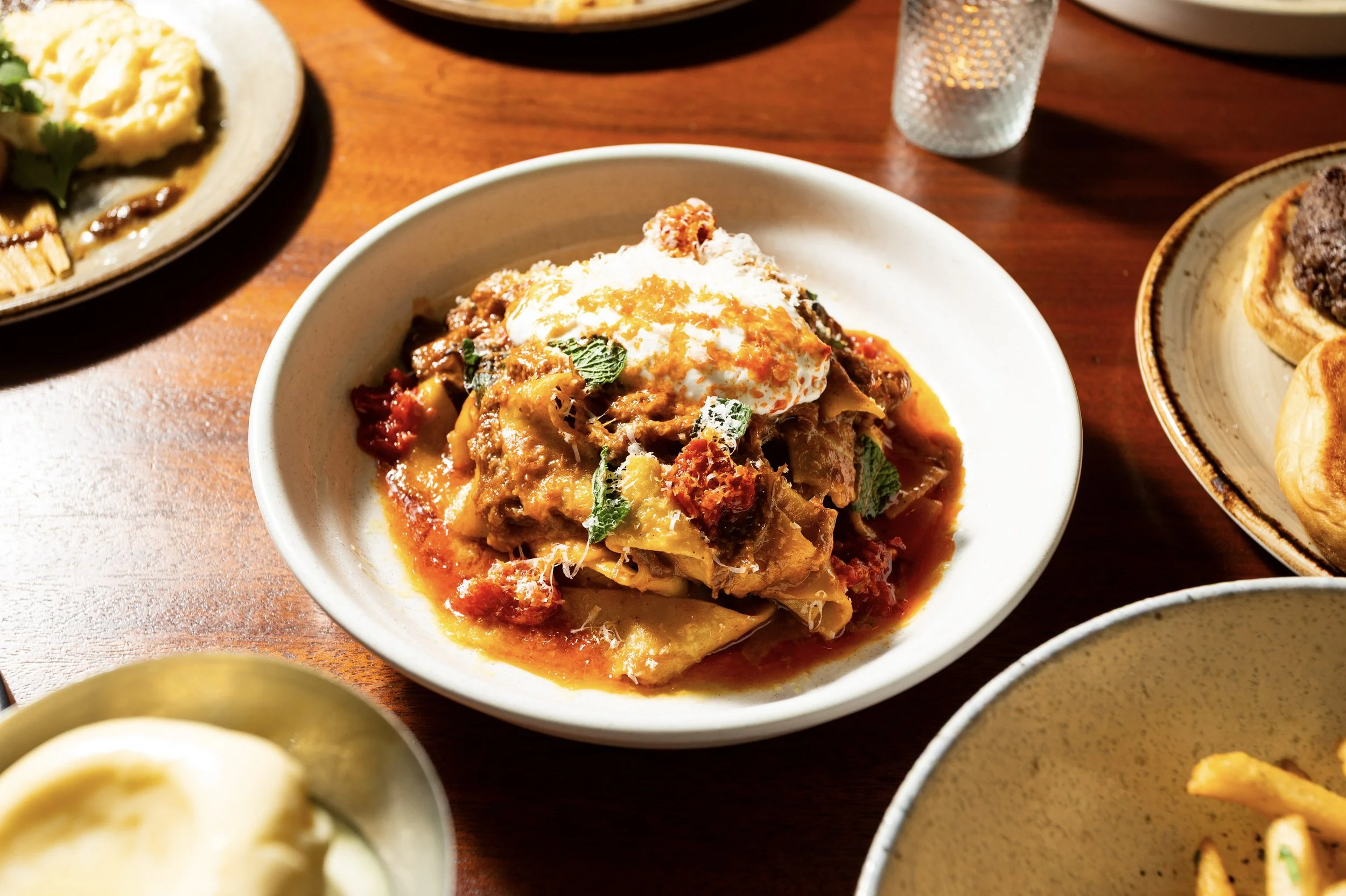 A white bowl of beef stew topped with a poached egg, garnished with herbs, and surrounded by other plates of food on a wooden table.