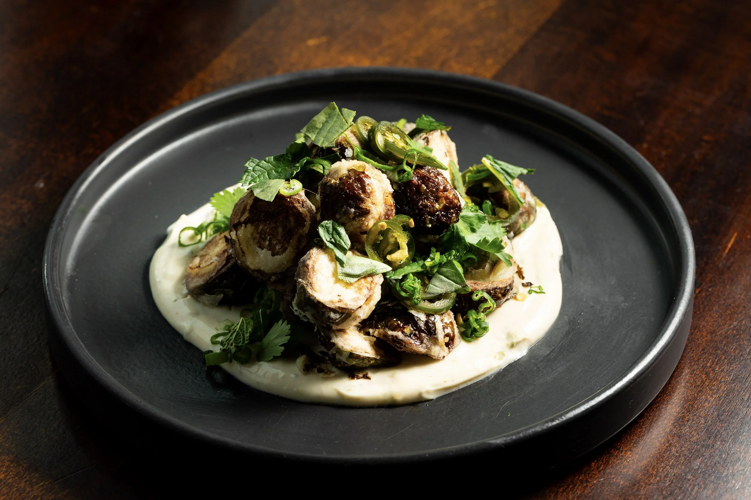 Dish of roasted eggplant stuffed with dark filling topped with chopped green herbs and sliced green chilies, served on a bed of white creamy sauce on a black plate.