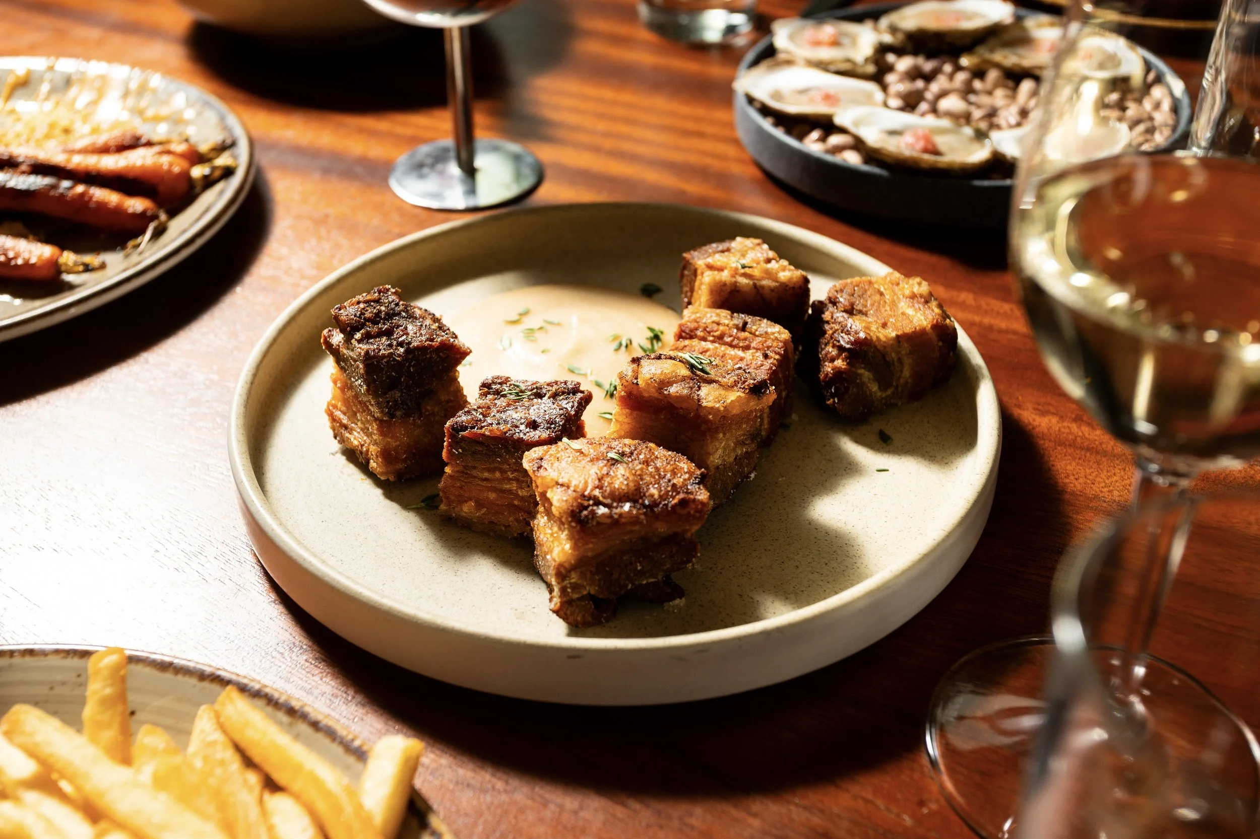Dishes of food on a wooden table, including fried pork belly with dipping sauce on a beige plate, fried potatoes, oysters, baked beans, and glasses of white wine.