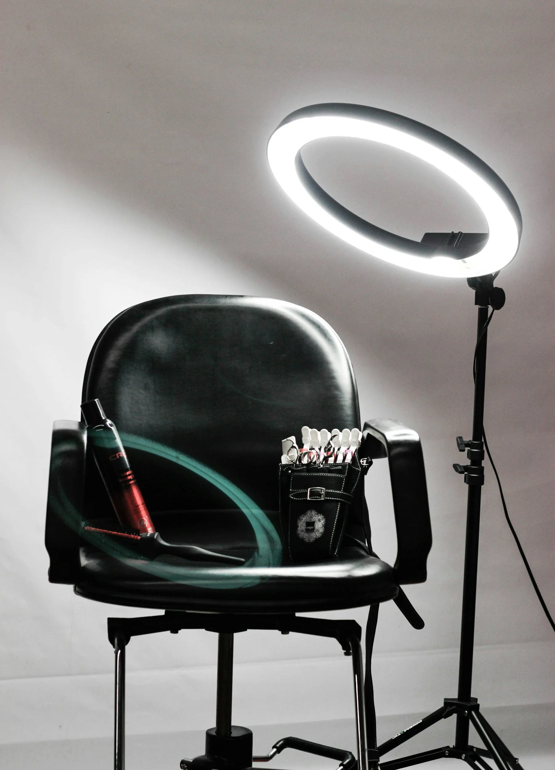 A black chair with a small black toolbag containing various makeup brushes and tools, a red cylindrical device, and a green item on the armrest. A ring light on a stand illuminates the scene against a plain white background.